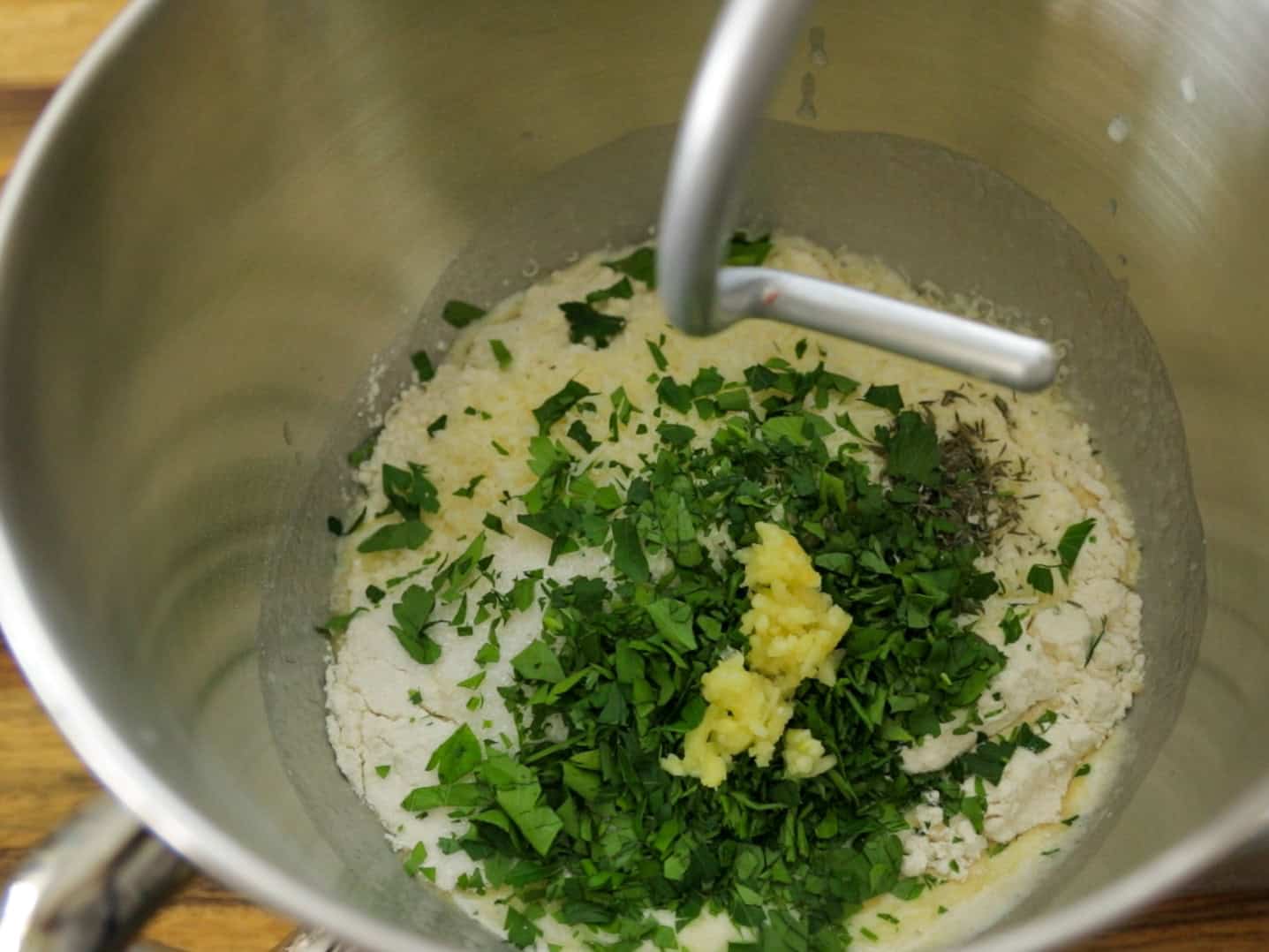A stand mixer bowl containing flour, chopped fresh herbs, and minced garlic, ready to be mixed with a dough hook attachment.