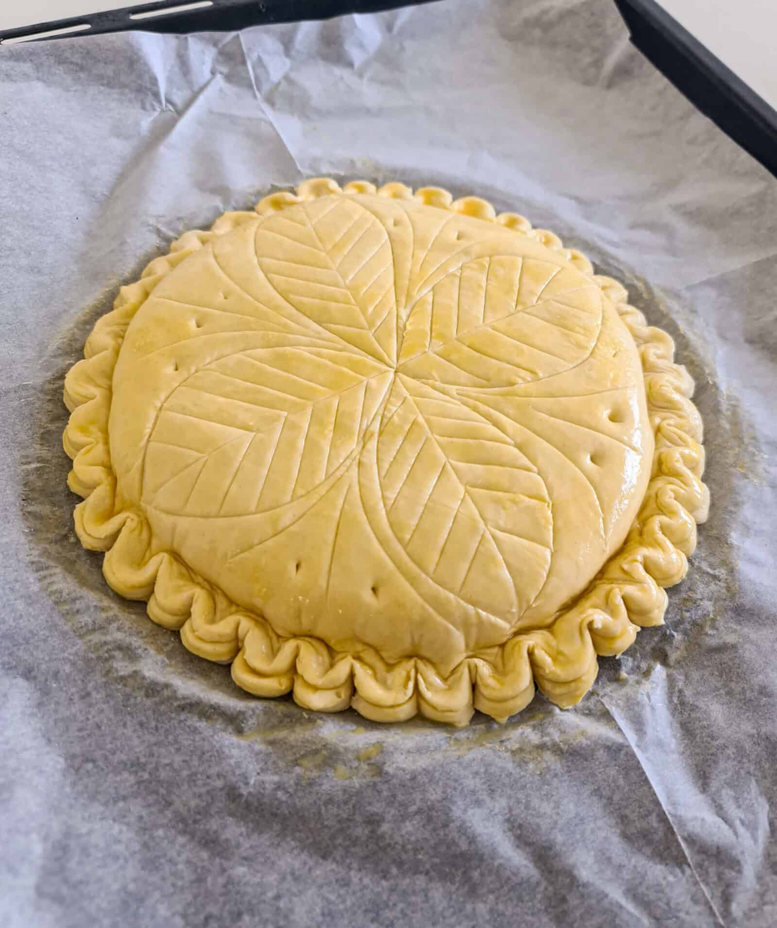 Unbaked galette des rois decorative leaf patterns and crimped edges, resting on parchment paper on a baking tray.