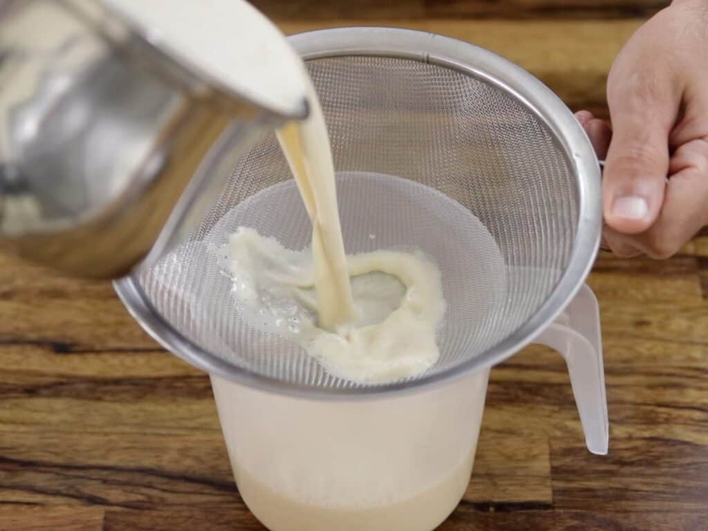 A person pours a creamy liquid from a metal container through a mesh strainer into a clear plastic measuring jug on a wooden surface.