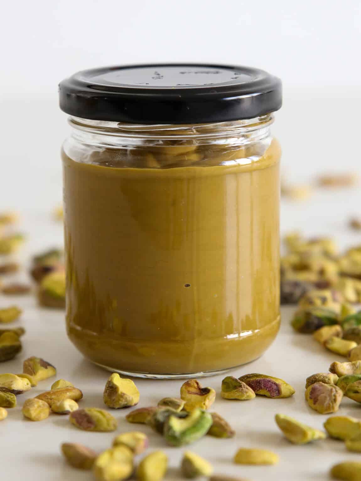 A glass jar filled with smooth pistachio paste, sealed with a black lid, surrounded by scattered shelled pistachios on a light surface.
