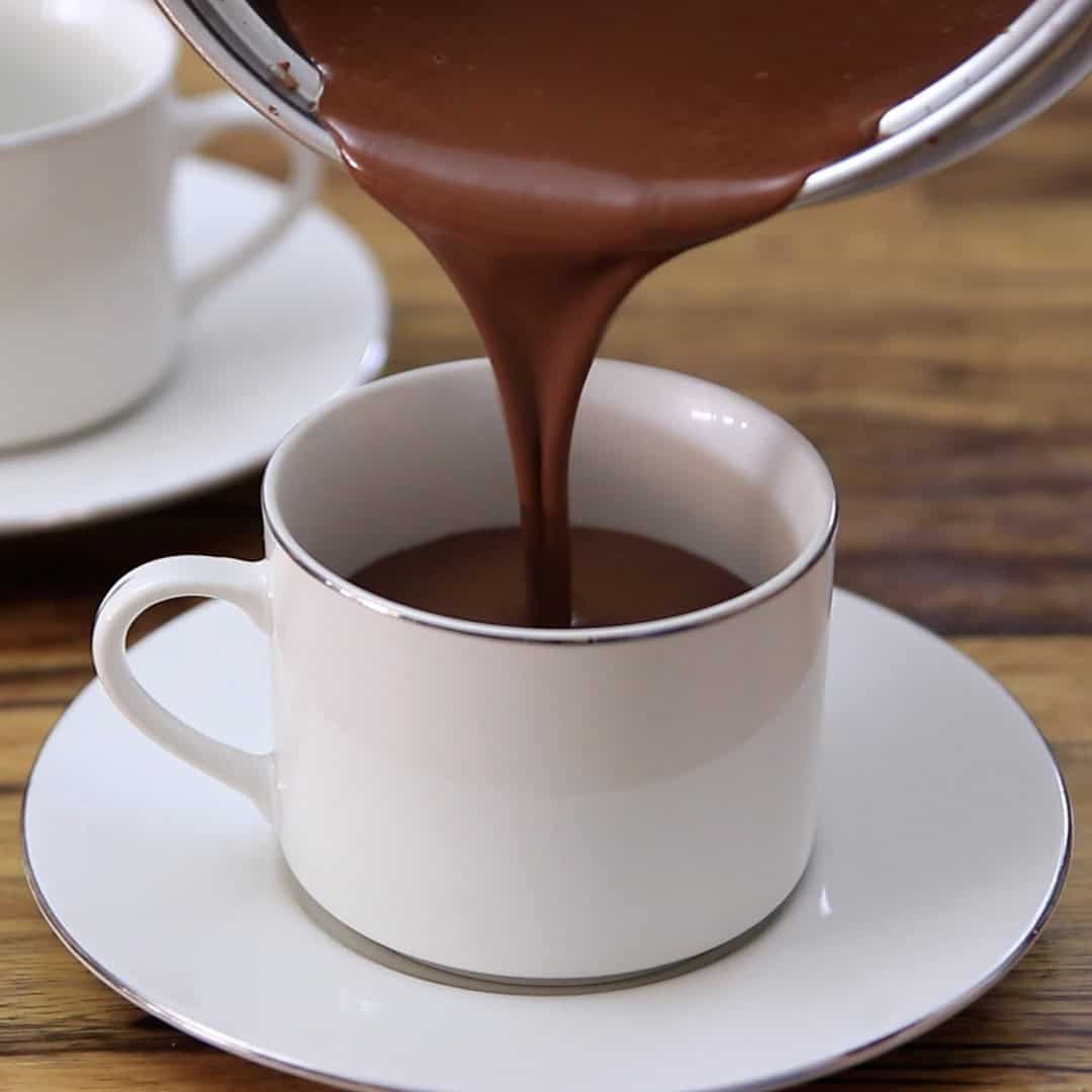 A thick stream of hot chocolate is being poured from a pot into a white cup on a matching saucer, placed on a wooden table. Another cup and saucer are visible in the background.