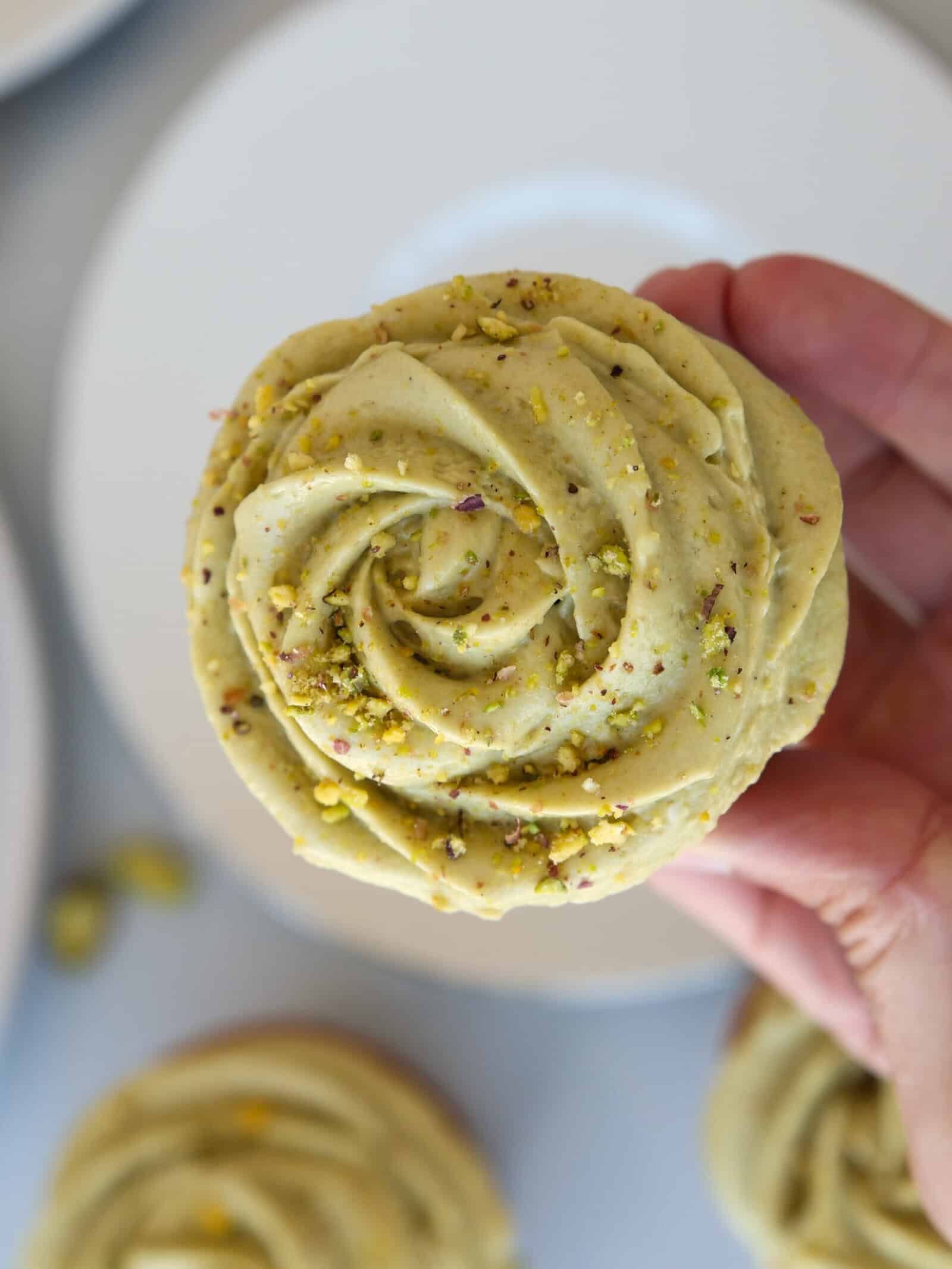 A hand holds a cupcake topped with creamy, pale green pistachio frosting swirled in a rose pattern and sprinkled with crushed pistachios. A white plate is seen in the background.