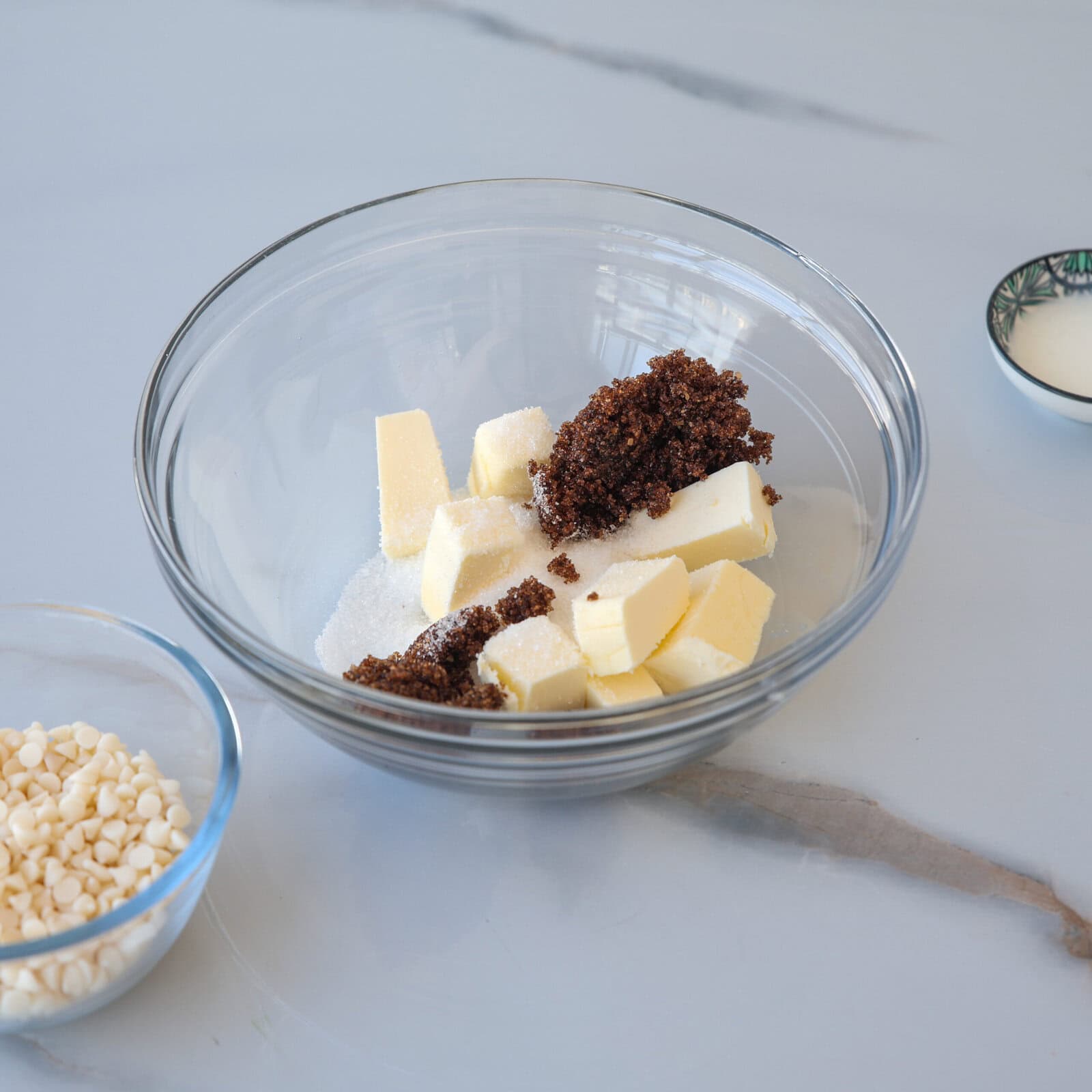 A glass bowl with chunks of butter, white granulated sugar, and brown sugar on a light gray surface. Nearby are a small bowl of white chocolate chips and a dish with liquid, possibly milk.