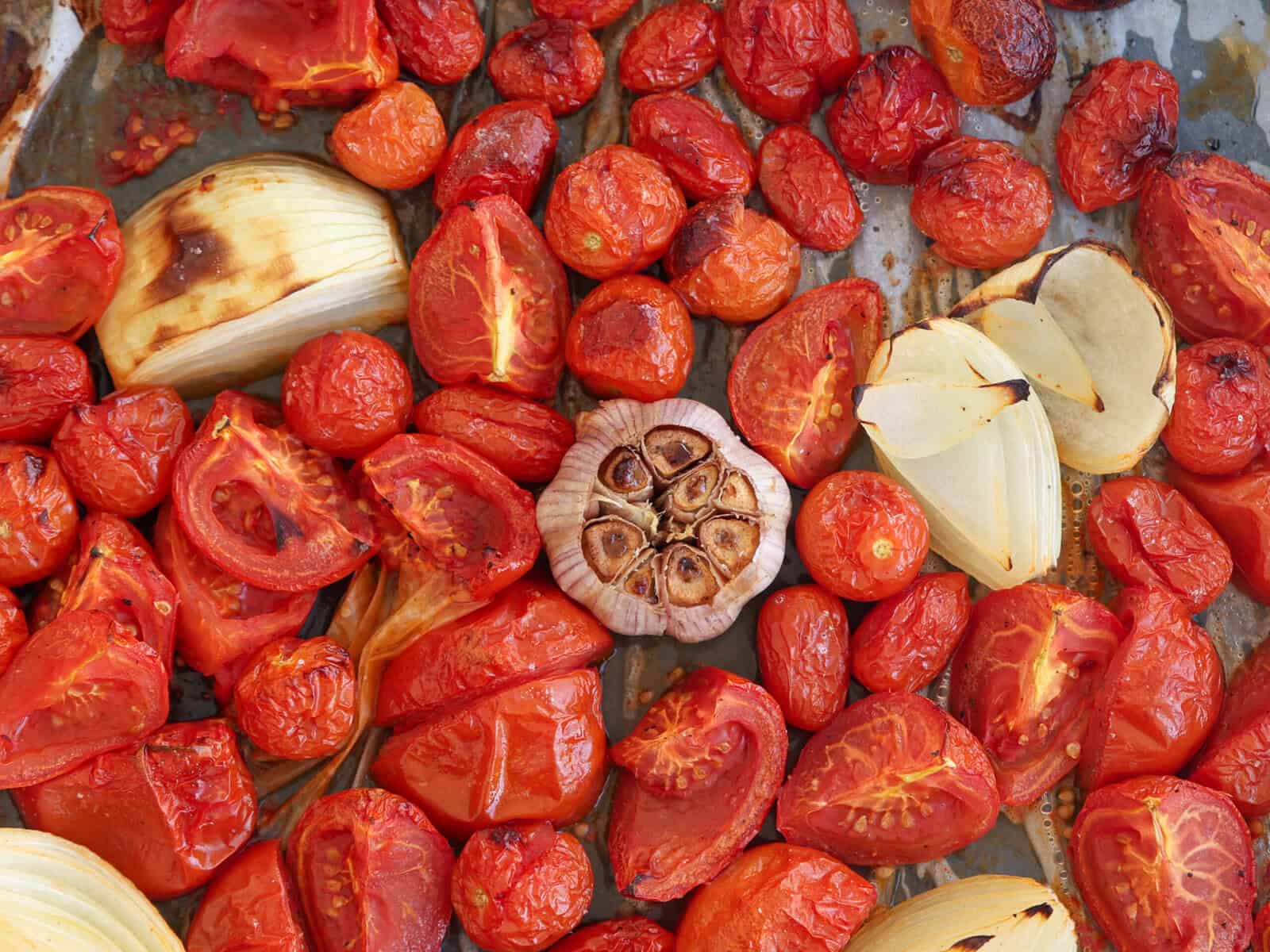 A baking tray with roasted tomatoes, cherry tomatoes, a halved garlic bulb, and caramelized onion wedges, all browned and slightly charred.