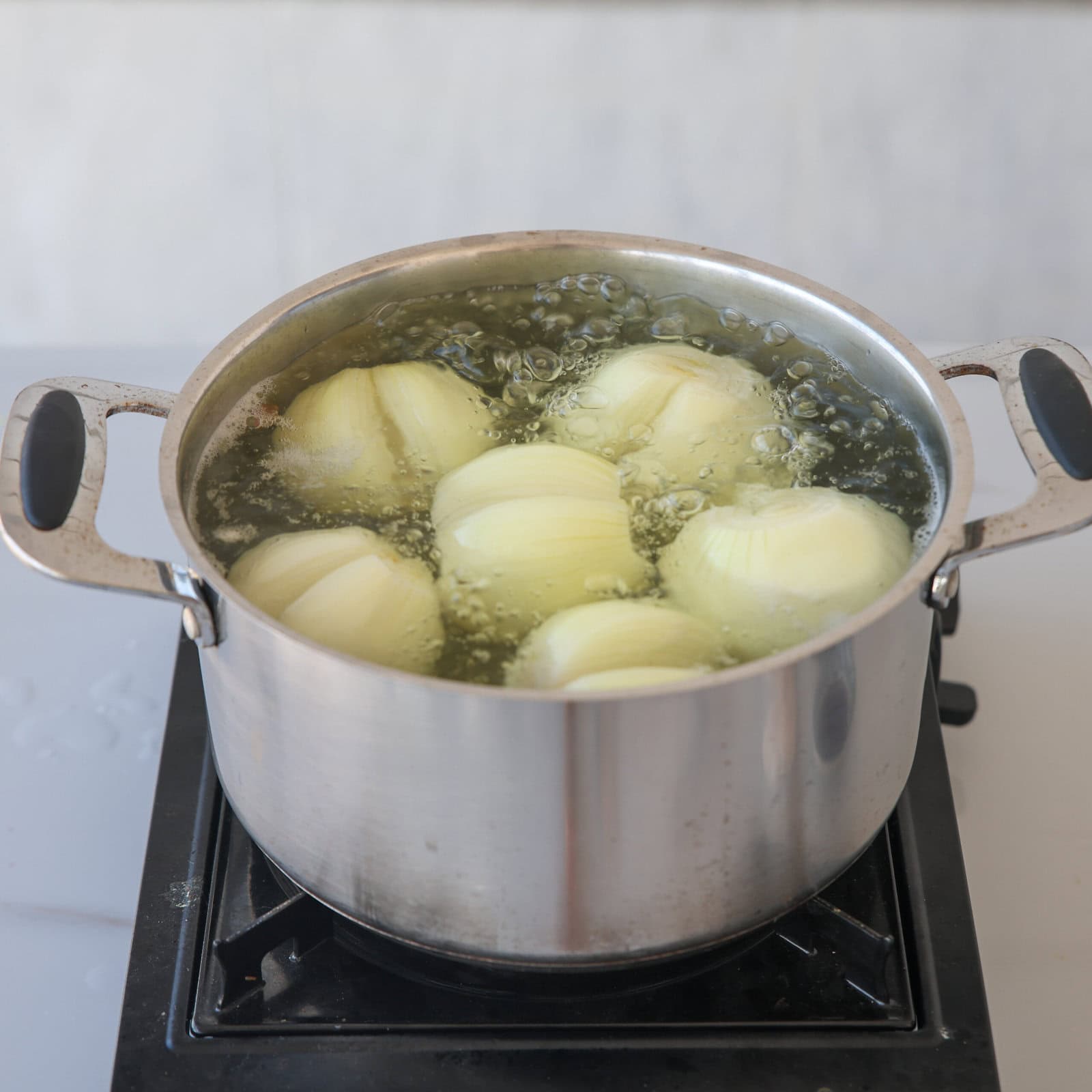 A stainless steel pot filled with peeled onions is boiling in water on a stovetop burner.