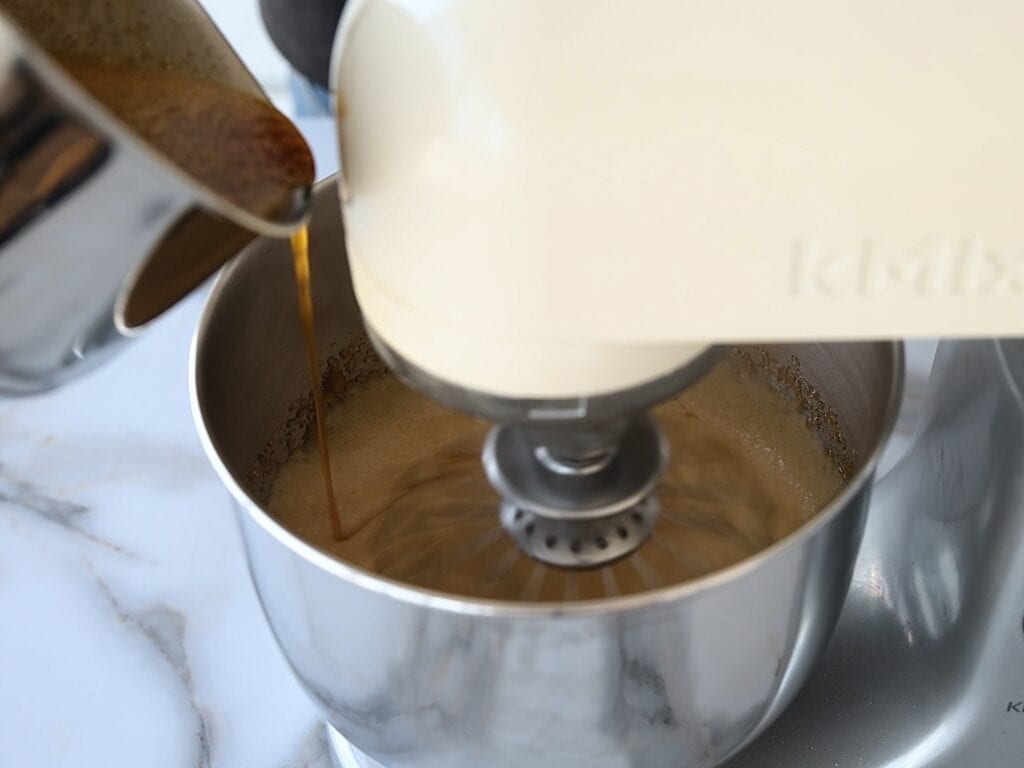 A close-up of a stand mixer whipping ingredients in a metal bowl while liquid is being poured in from a saucepan on the left side. The scene is set on a white marble countertop.