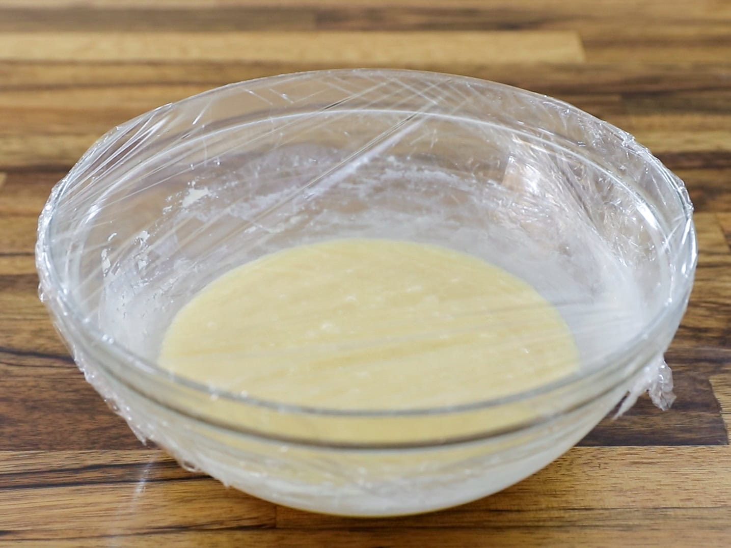 A glass bowl with dough covered in plastic wrap sits on a wooden surface, ready for proofing.