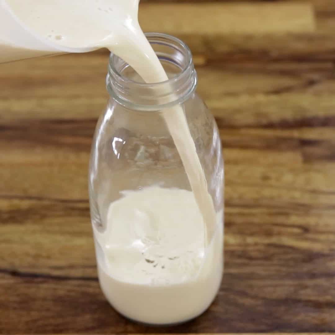 A creamy eggnog is being poured from a pitcher into a clear glass bottle on a wooden surface.