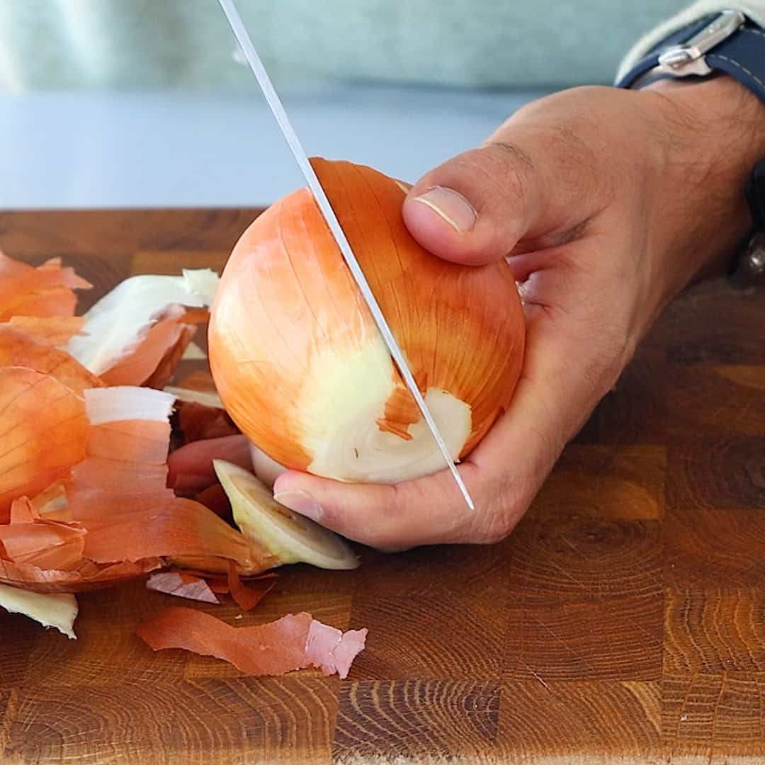 A person slicing an onion in half on a wooden cutting board, with onion skins and pieces scattered around.
