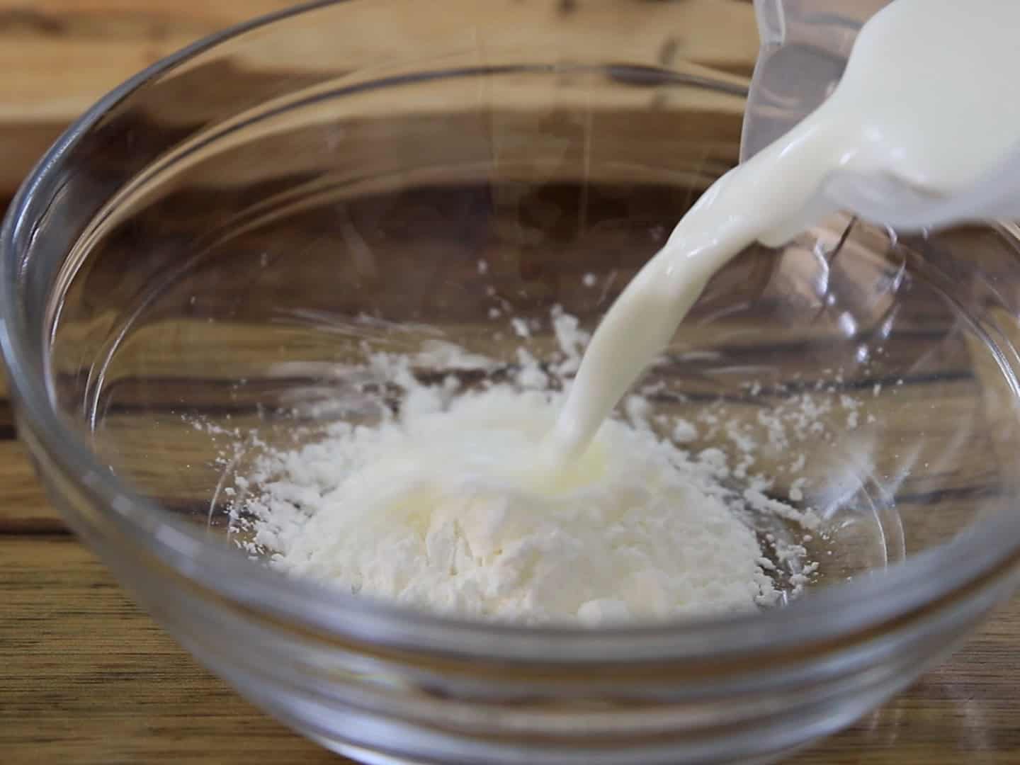 A clear glass bowl containing white powder is on a wooden surface. Milk is being poured from a container into the bowl, mixing with the powder.