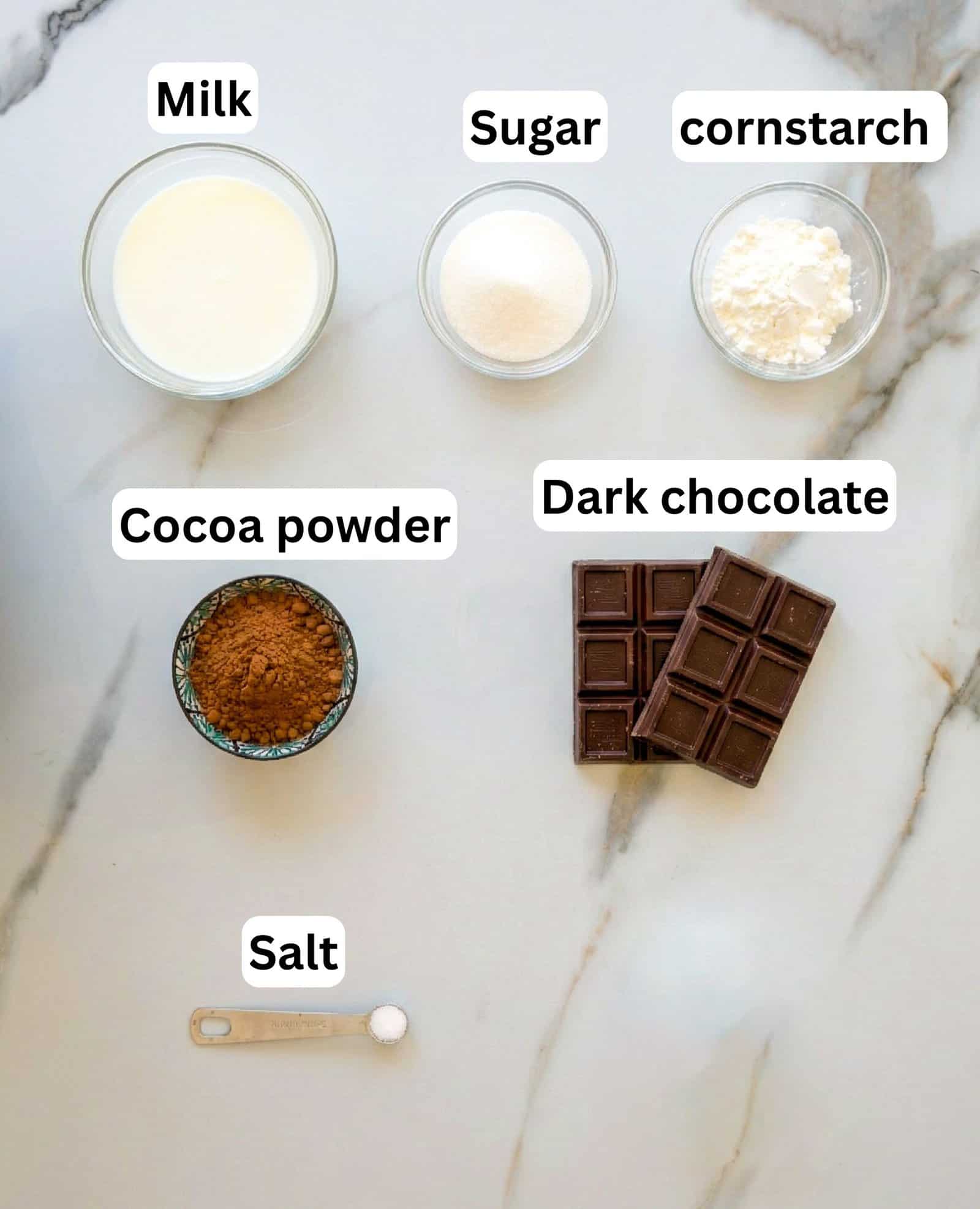 Overhead view of labeled ingredients on a light surface: a bowl of milk, a bowl of sugar, a bowl of cornstarch, a bowl of cocoa powder, two bars of dark chocolate, and a teaspoon of salt.