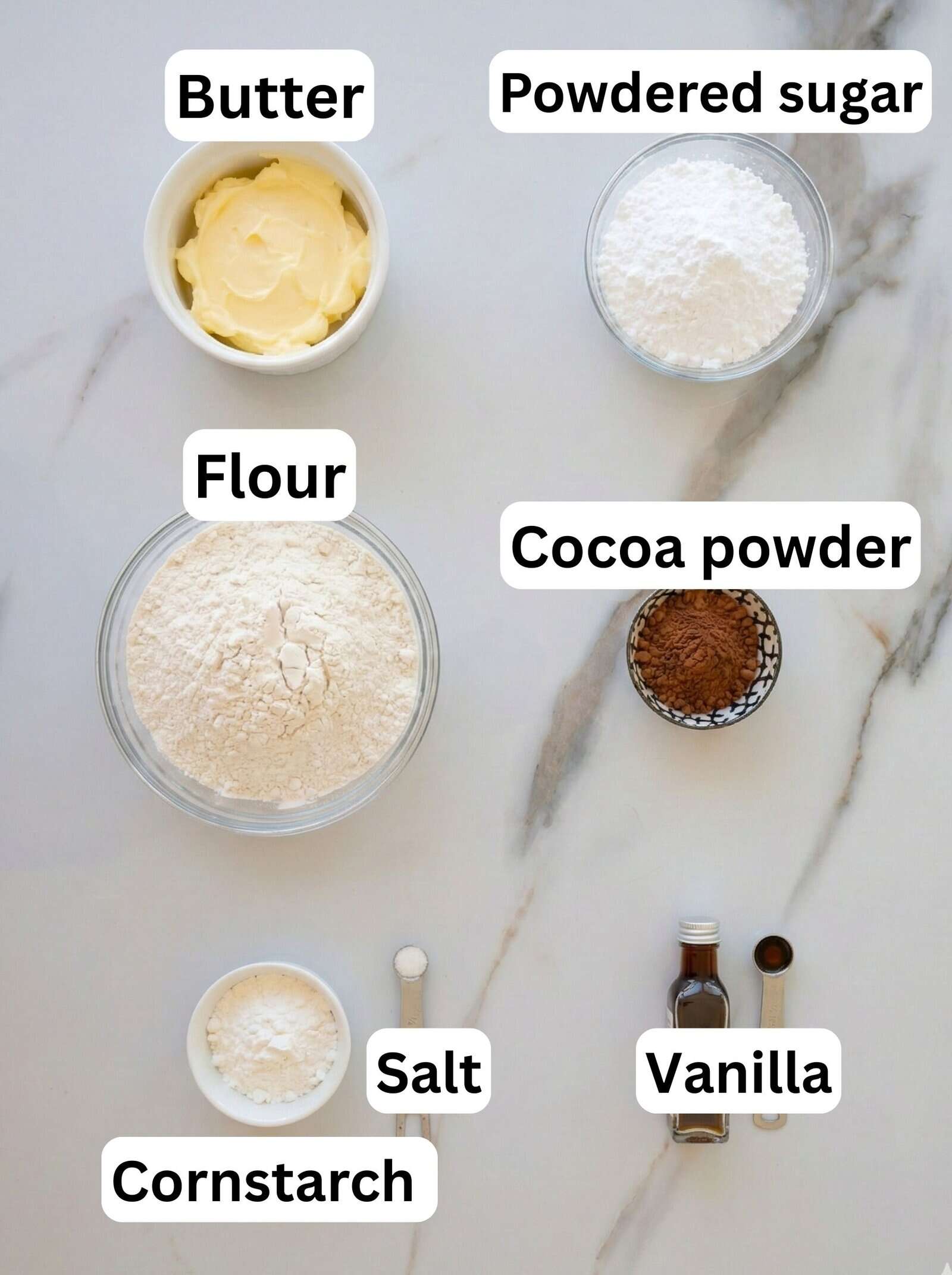 Baking ingredients on a marble surface: bowls of butter, powdered sugar, flour, cocoa powder, and cornstarch, plus small amounts of salt and vanilla extract, each labeled with text.