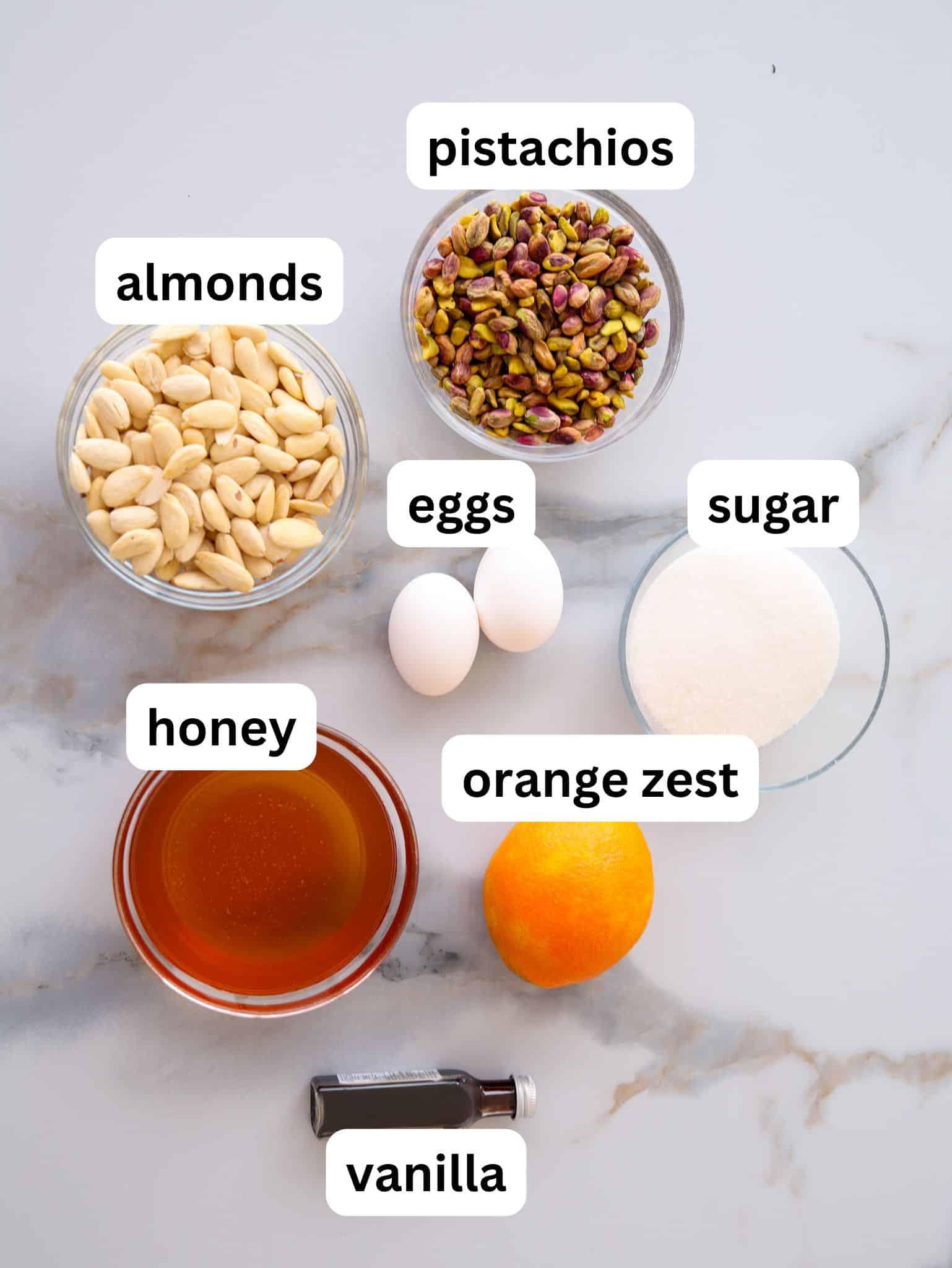 A marble countertop displays labeled ingredients in bowls: almonds, pistachios, eggs, sugar, honey, an orange for zest, and a bottle of vanilla extract.
