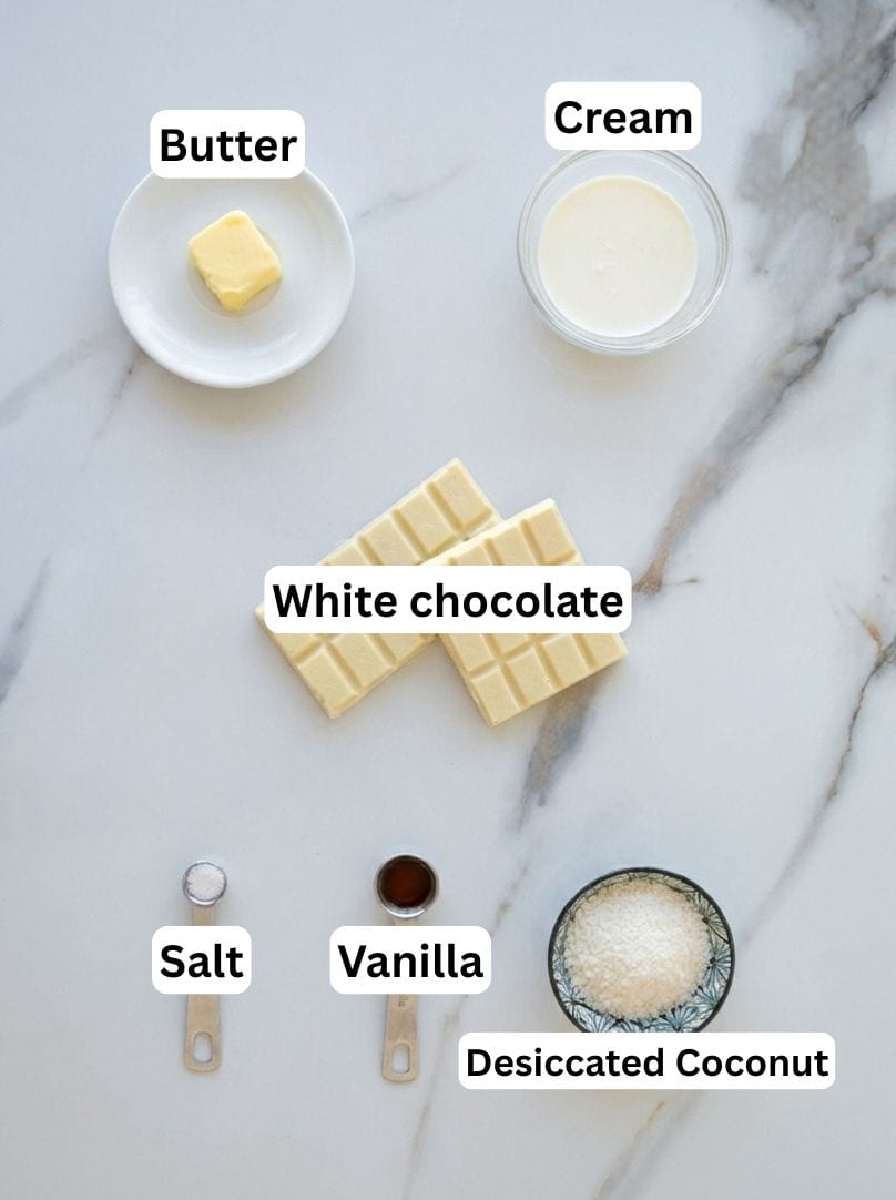 A flat lay of baking ingredients on a white surface, including butter on a plate, a bowl of cream, white chocolate bars, a small bowl of desiccated coconut, a spoon of salt, and a spoon of vanilla extract. Each is labeled.