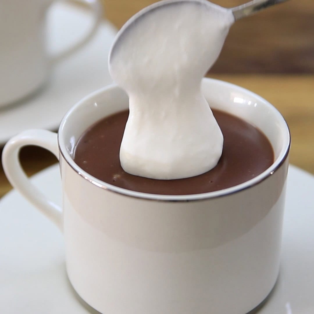 A spoon is lifting a dollop of whipped cream above a cup filled with hot chocolate, with another cup blurred in the background.