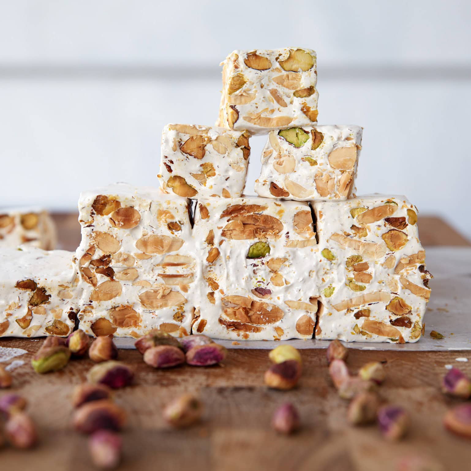 Pieces of nut-filled nougat stacked in a pyramid on a wooden surface, with whole pistachios scattered in front. The nougat is white and studded with various nuts.