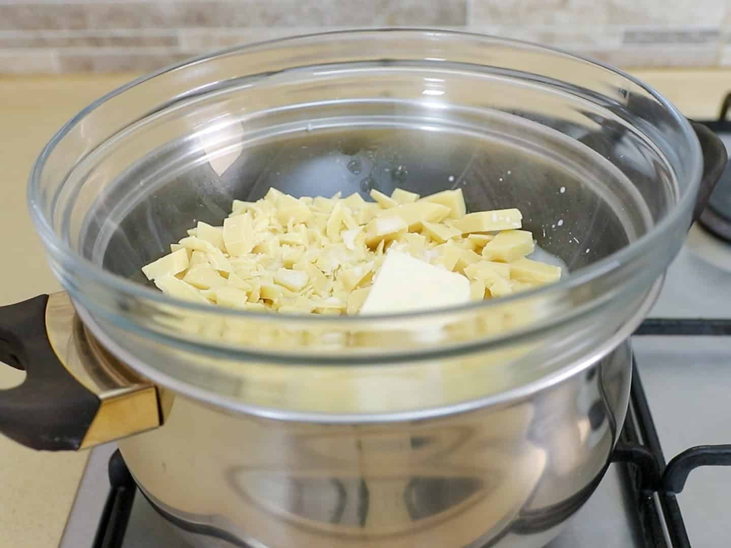 A glass bowl with chopped white chocolate sits on top of a pot of hot water on a stovetop, forming a double boiler for melting the chocolate.