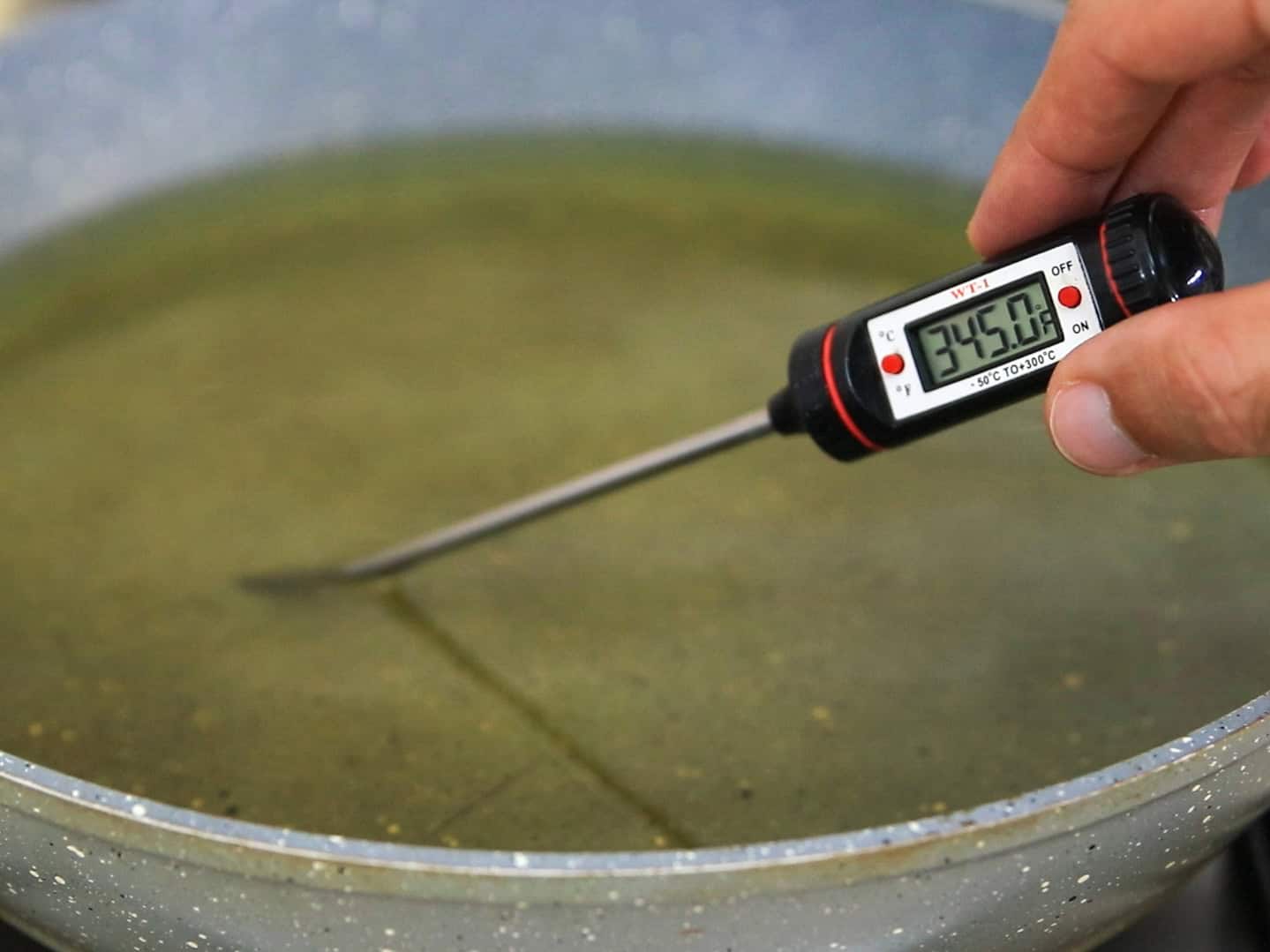 A hand holds a digital thermometer, measuring the temperature of oil in a frying pan. The thermometer displays 345.0°F.