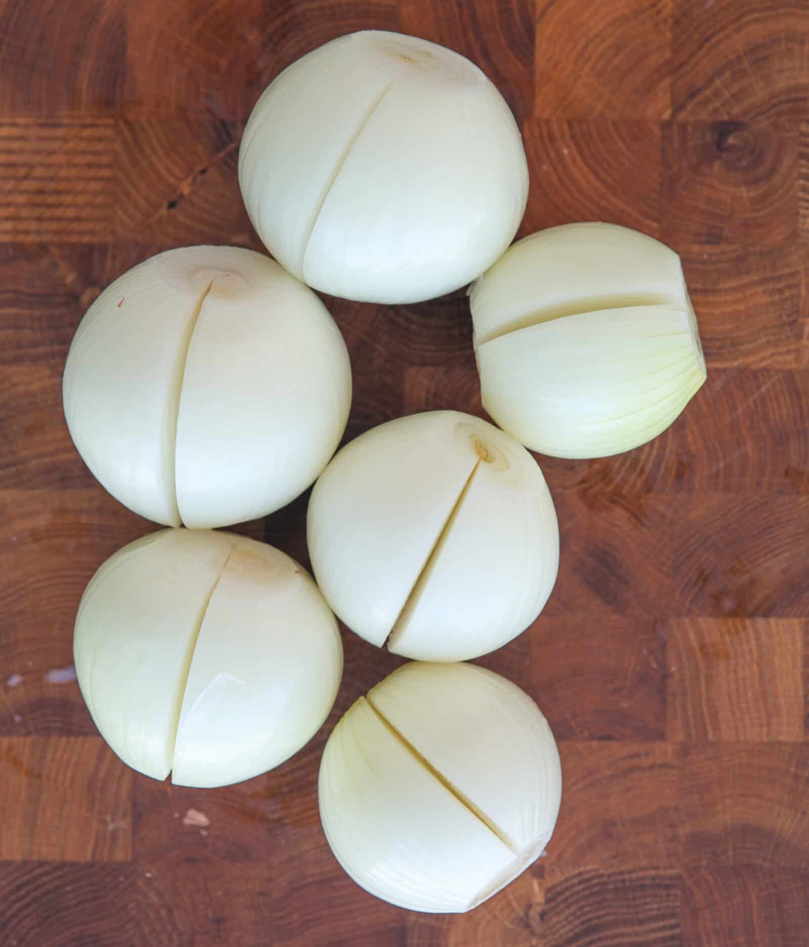 Six peeled onions, each sliced halfway through the center, are arranged on a wooden cutting board with a checkered pattern.