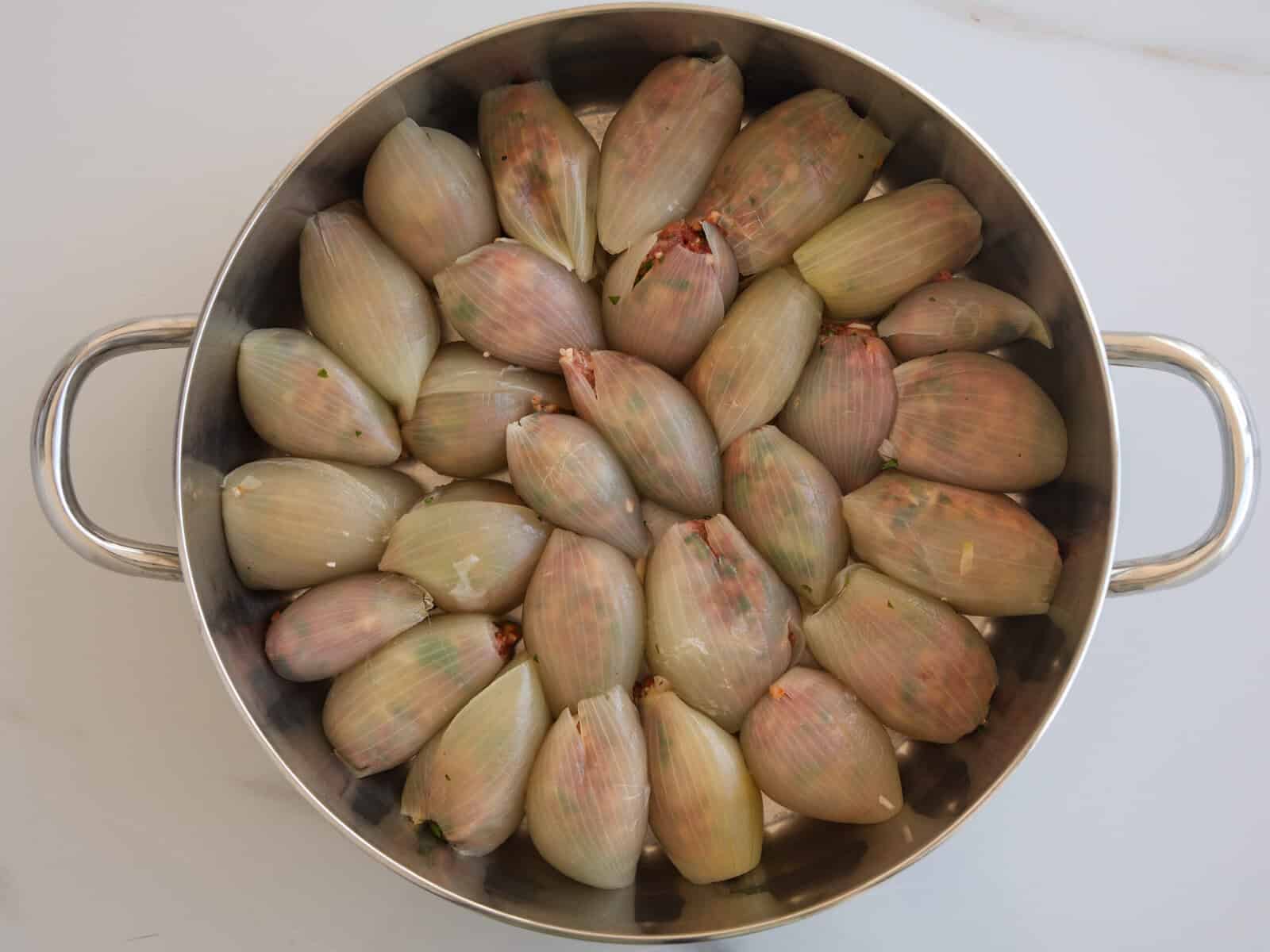 A large metal pot filled with stuffed onion shells, neatly arranged in a circular pattern before cooking. The onions appear tightly packed and lightly tinged with green and pink hues.