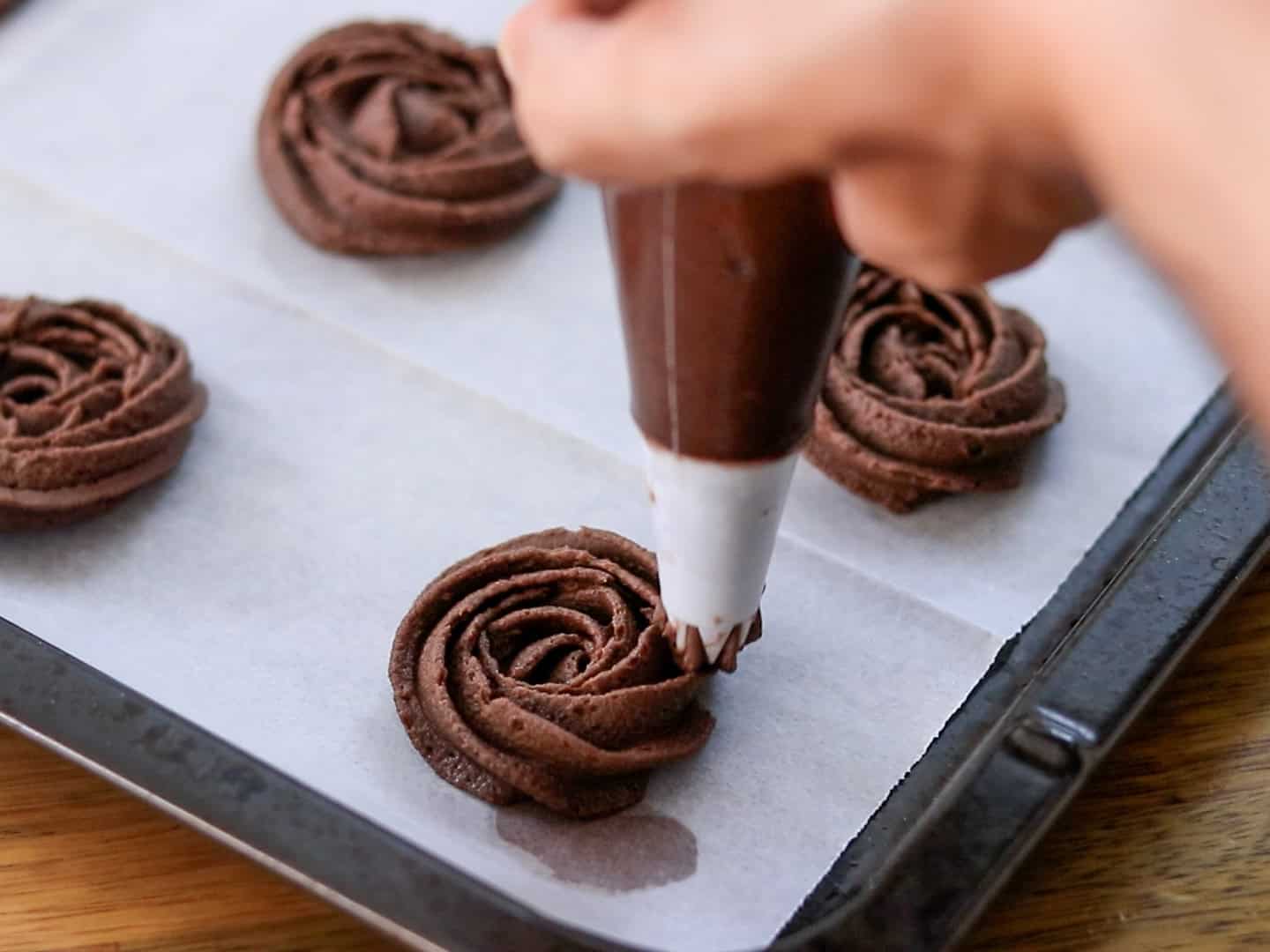 A hand uses a piping bag to form chocolate cookie dough into swirled shapes on a parchment-lined baking tray.