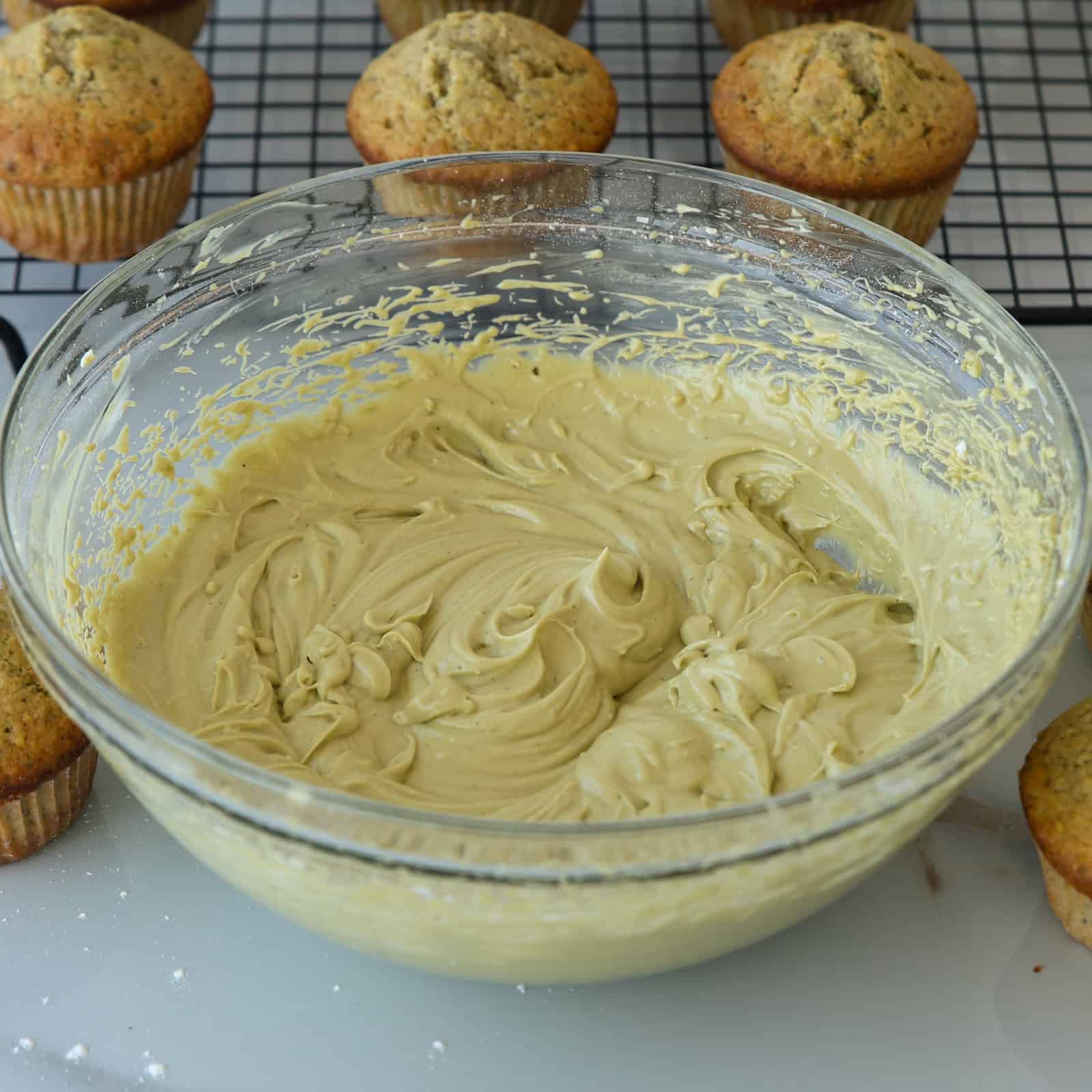 A glass bowl filled with creamy pistachio cream cheese frosting sits on a countertop surrounded by freshly baked muffins and a cooling rack in the background.