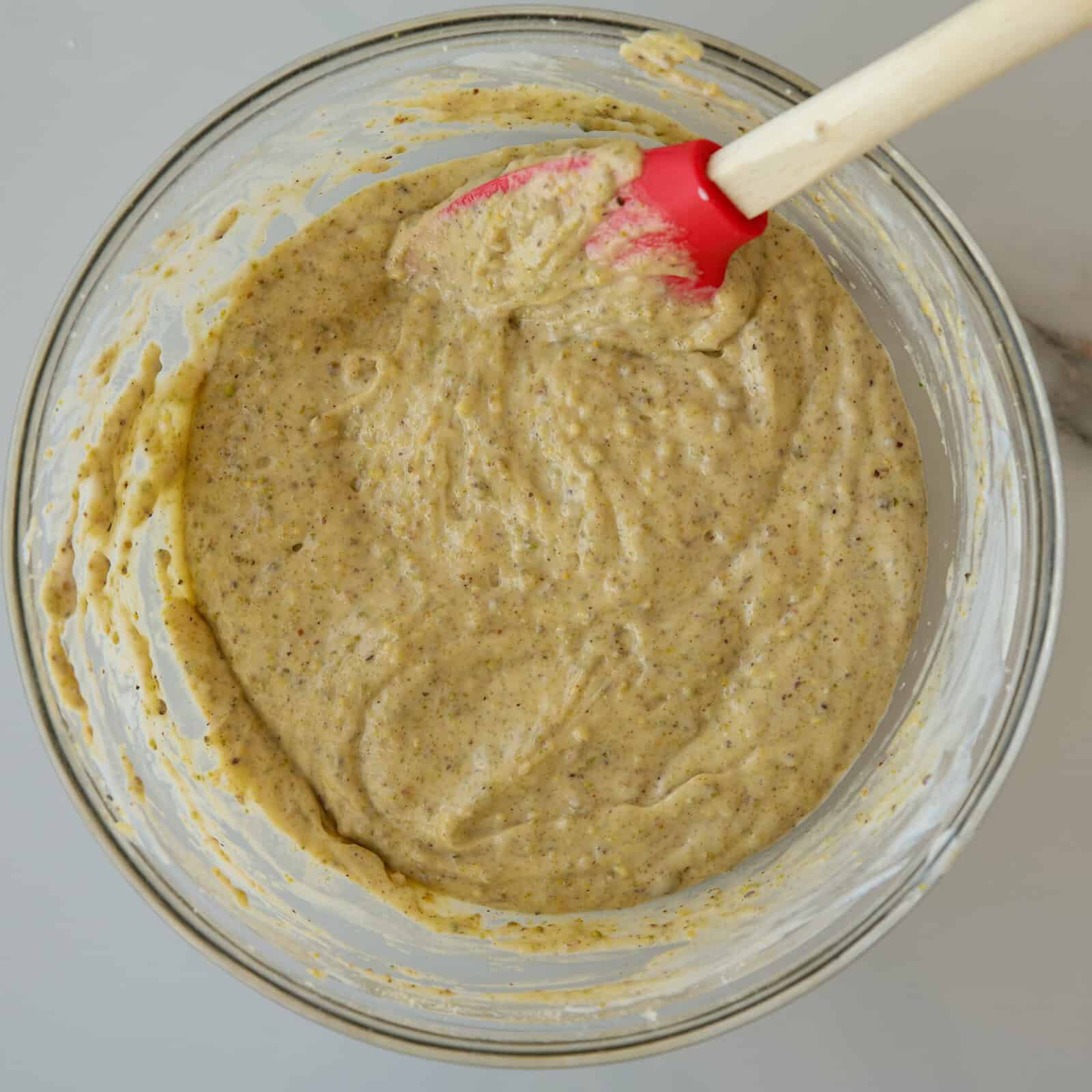 A glass bowl filled with thick, yellowish greem pistachio batter speckled with dark flecks, being mixed with a red and white spatula. The bowl sits on a light-colored surface.