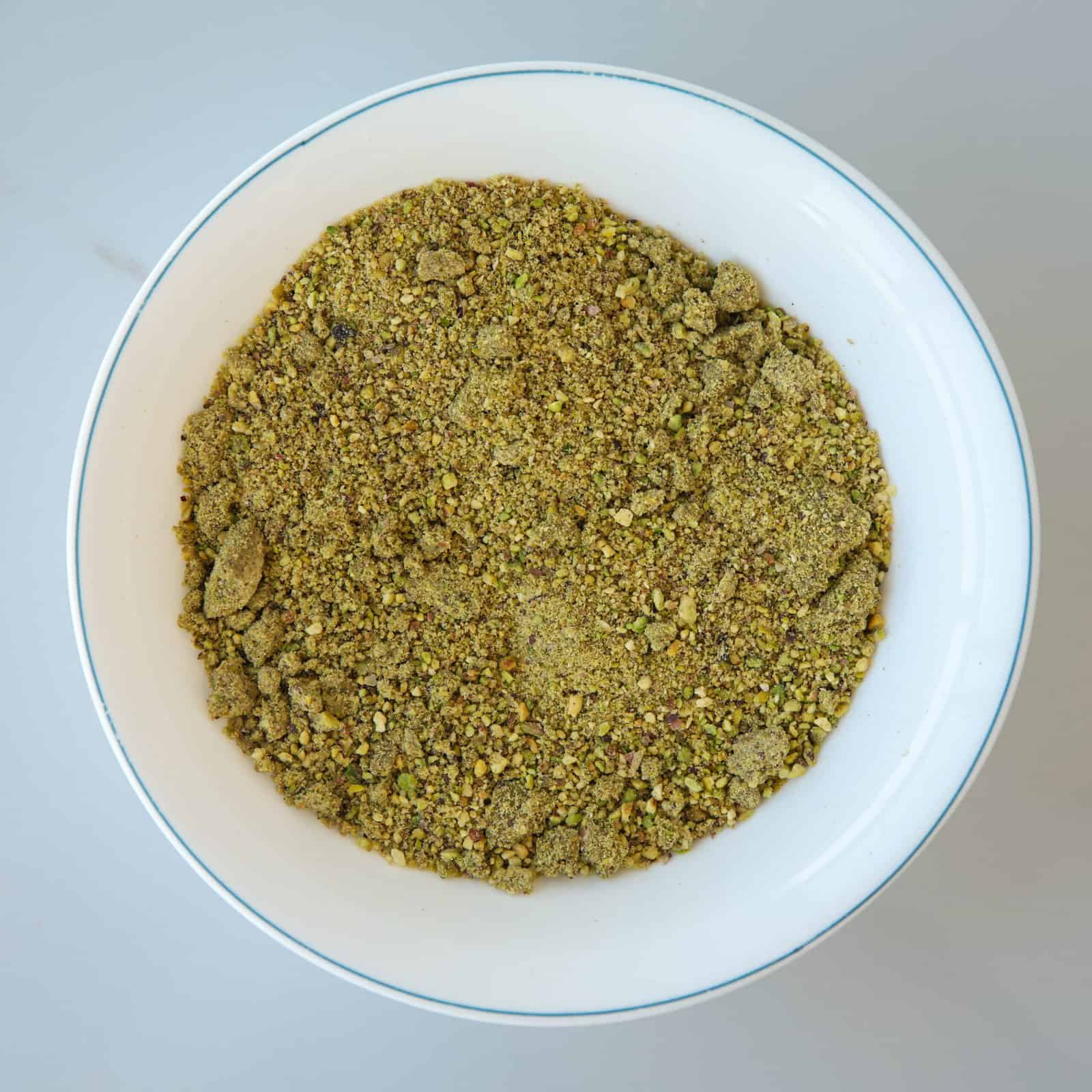 A white bowl filled with ground pistachios (pistachio flour), placed on a light gray surface.