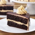 A slice of chocolate layer cake with light peanut butter frosting between the layers and on top, garnished with a swirl of frosting, served on a white plate.