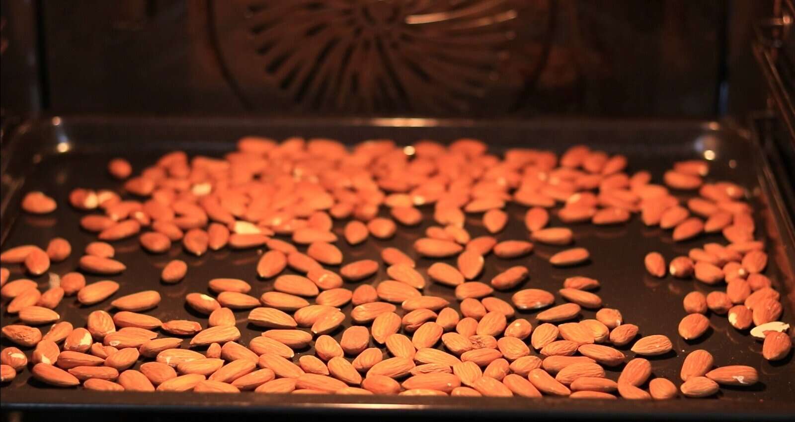 A baking tray filled with raw almonds is placed inside an oven, ready for roasting. The oven's interior and the scattered almonds are clearly visible.