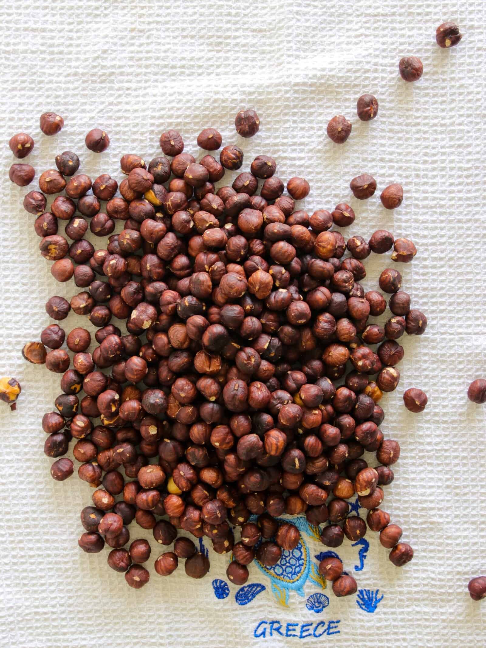 A pile of roasted hazelnuts scattered on a textured white cloth with "GREECE" and a blue and yellow design printed near the bottom edge.