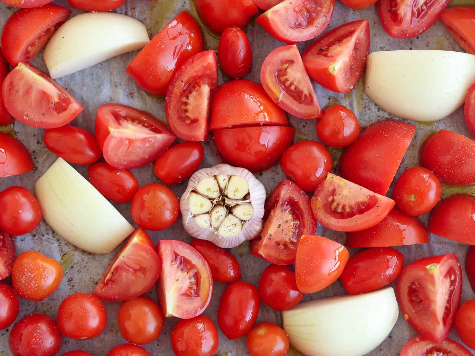 A variety of cut tomatoes, whole cherry tomatoes, quartered onions, and a halved garlic bulb are arranged on a baking sheet, ready for roasting.