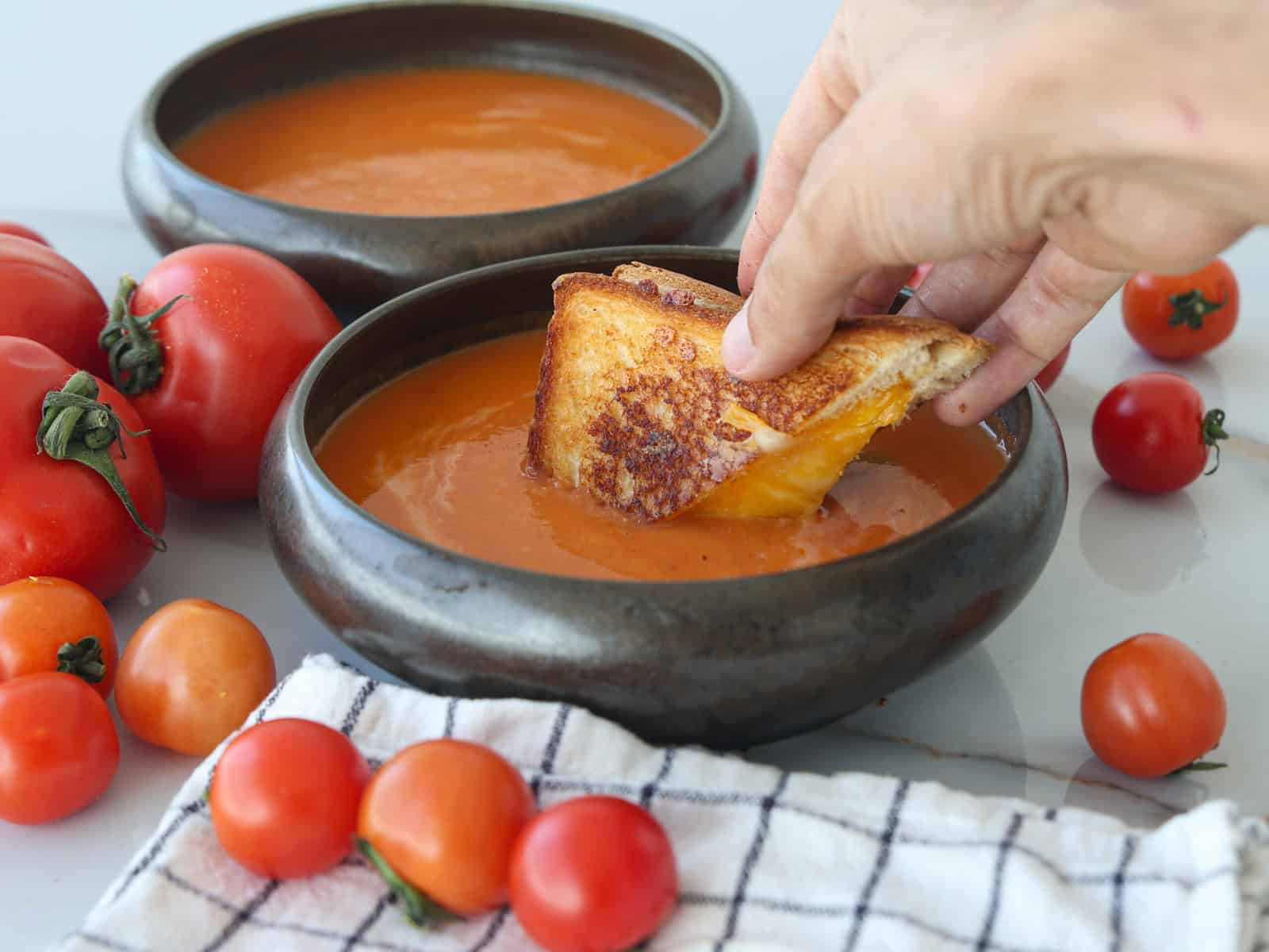 A hand dips a grilled cheese sandwich into a bowl of tomato soup, surrounded by fresh tomatoes and another bowl of soup, with a checkered cloth nearby.