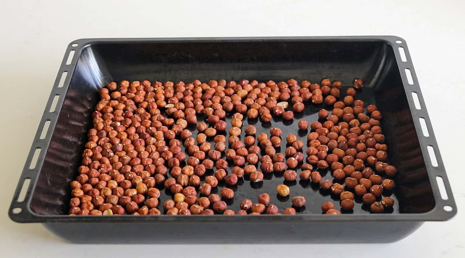 A black rectangular baking tray with a single layer of raw hazelnuts scattered unevenly across its surface, placed on a white countertop.