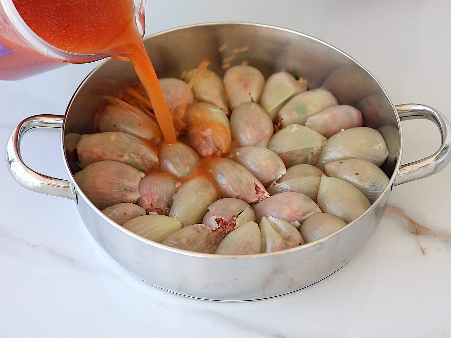A pot filled with stuffed onions arranged in a circle, with tomato sauce being poured over them from a measuring cup. The scene is set on a white countertop.