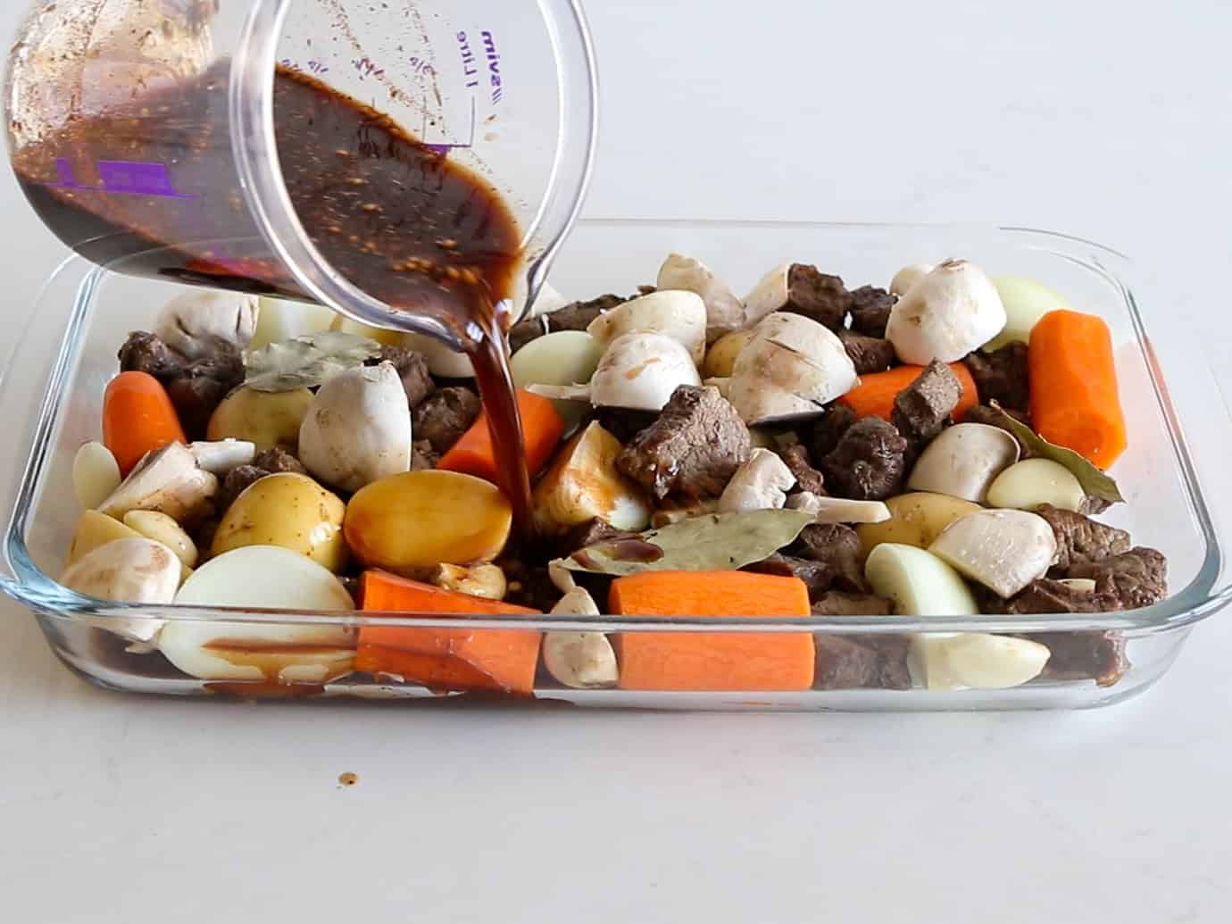 A glass baking dish filled with raw beef chunks, carrots, potatoes, onions, mushrooms, and bay leaves is being topped with a dark marinade poured from a measuring cup.
