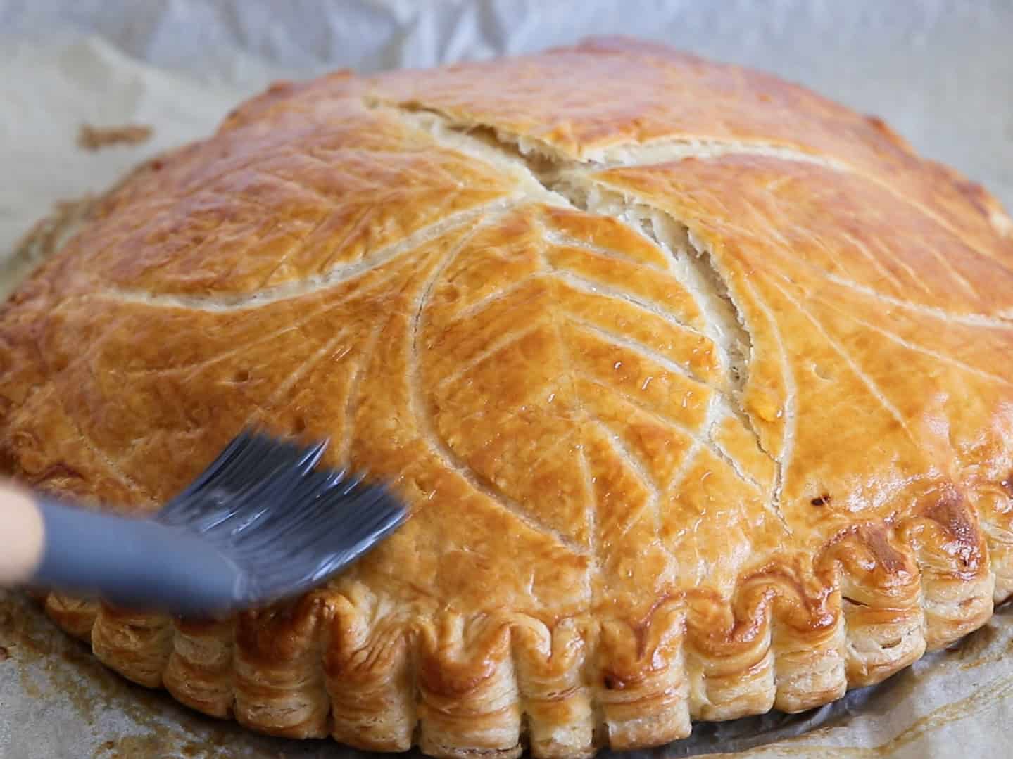 A close-up of a golden-brown round pastry with decorative leaf patterns on top, being brushed with a shiny glaze. The pie sits on baking parchment.