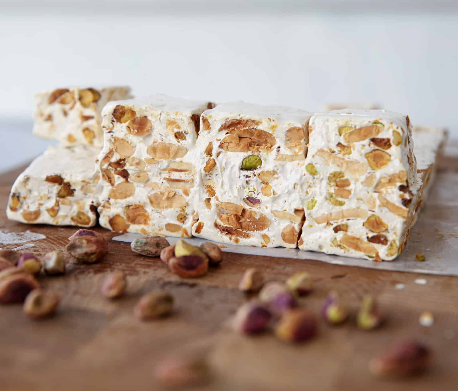 Blocks of torrone filled with assorted nuts, including pistachios and almonds, are stacked on a wooden surface with scattered whole nuts in the foreground.