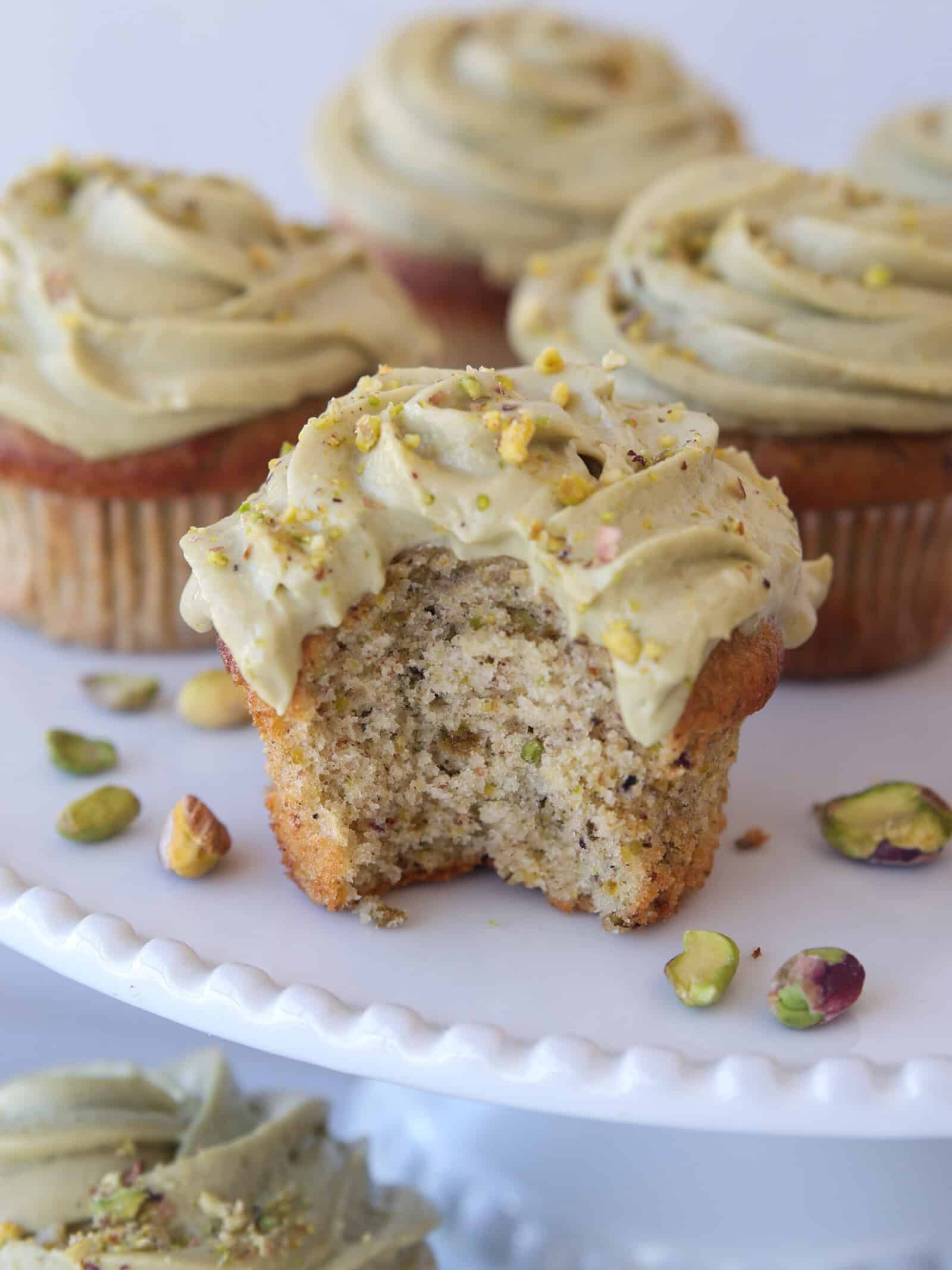 A pistachio cupcake with green frosting and chopped pistachios sits on a white plate, with a bite taken out, revealing a moist interior. More frosted cupcakes and scattered pistachios are in the background.
