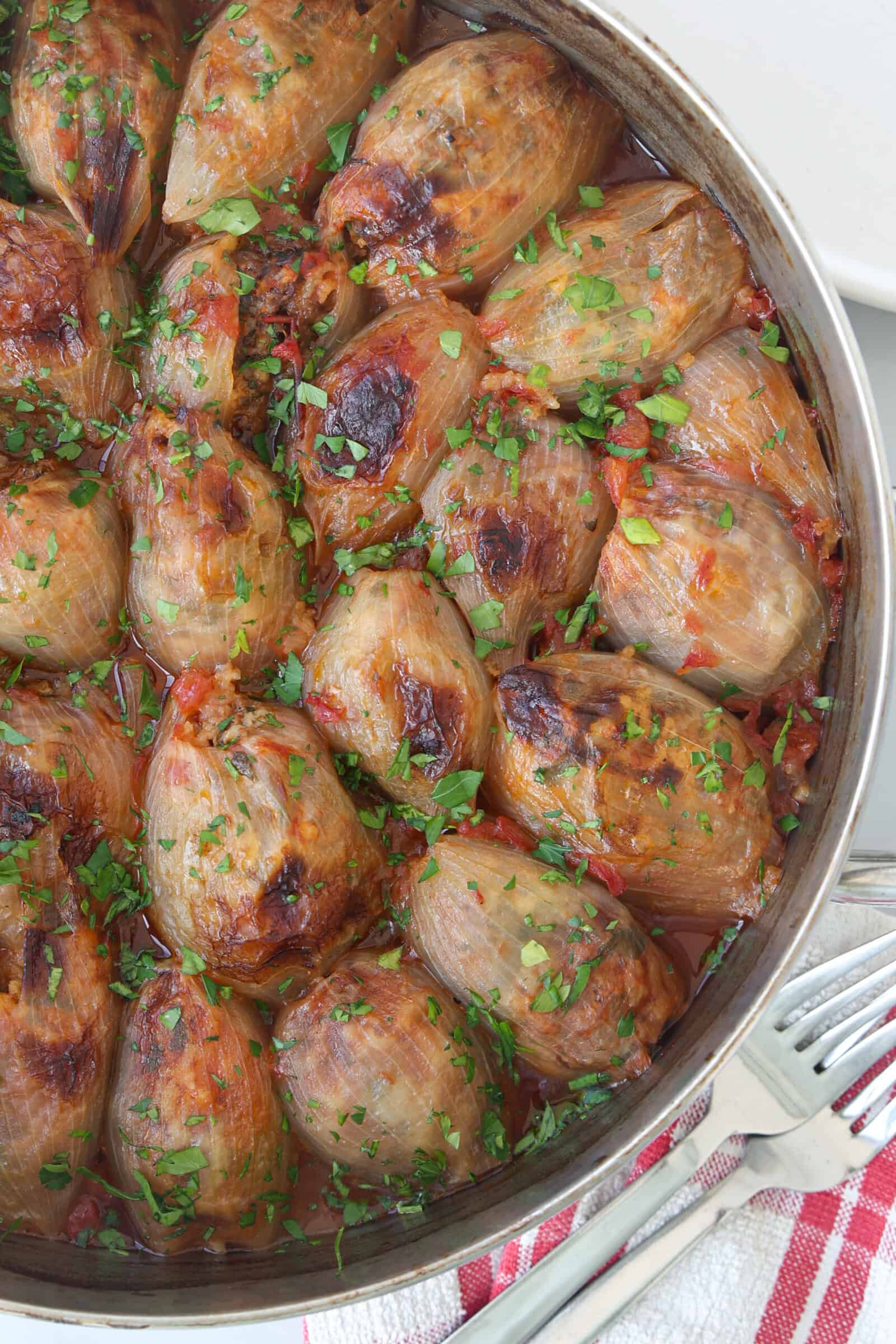 A pot filled with cooked stuffed onions, garnished with chopped parsley and herbs, in a tomato-based sauce. A red-and-white checkered towel with utensils is beside the pot.