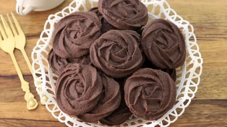 A white lace-patterned plate holds a pile of chocolate butter cookies shaped like roses. Two gold forks rest on the wooden table beside the plate.