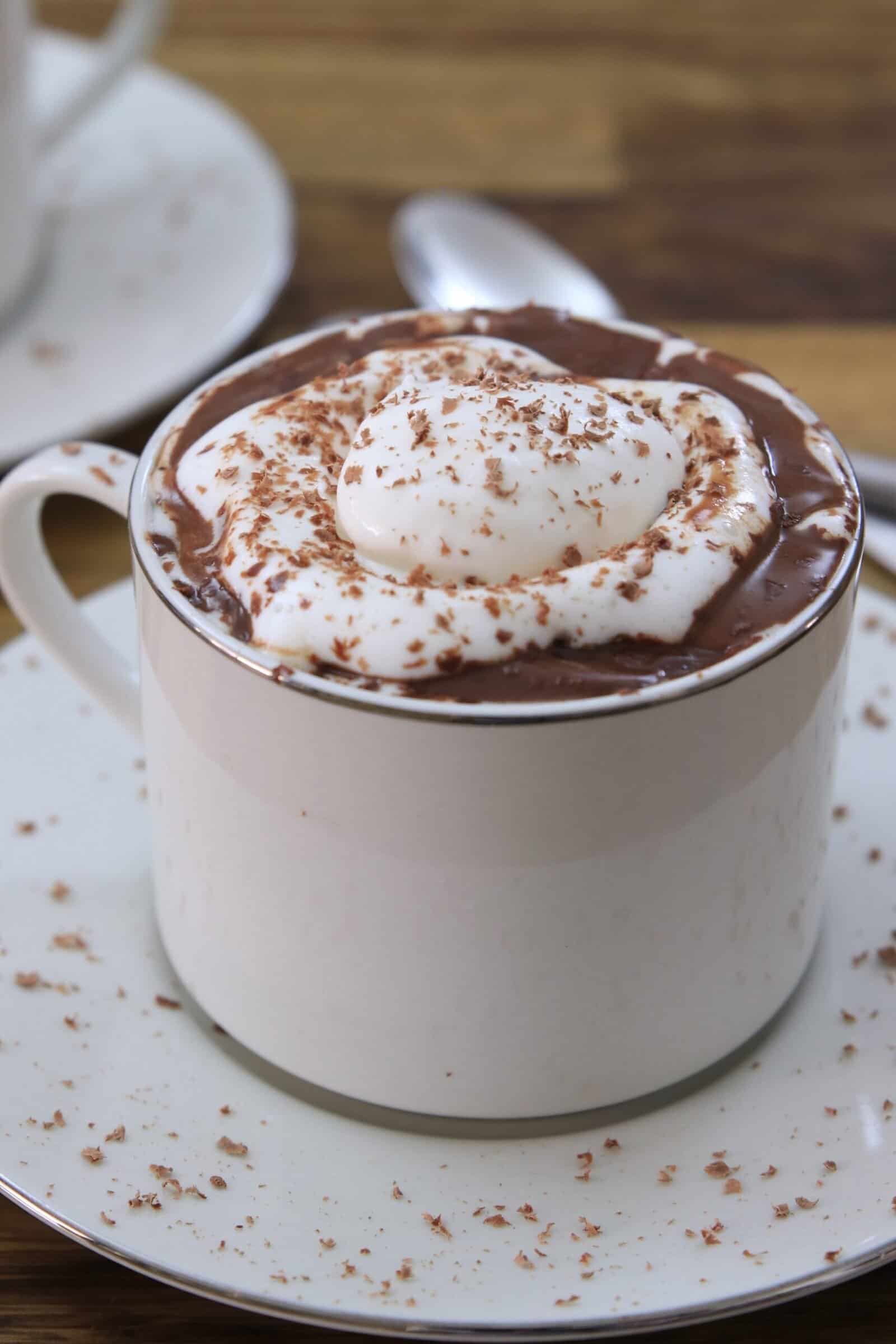 A white cup filled with thick italian hot chocolate topped with whipped cream and chocolate shavings, sitting on a matching saucer with a spoon in the background.