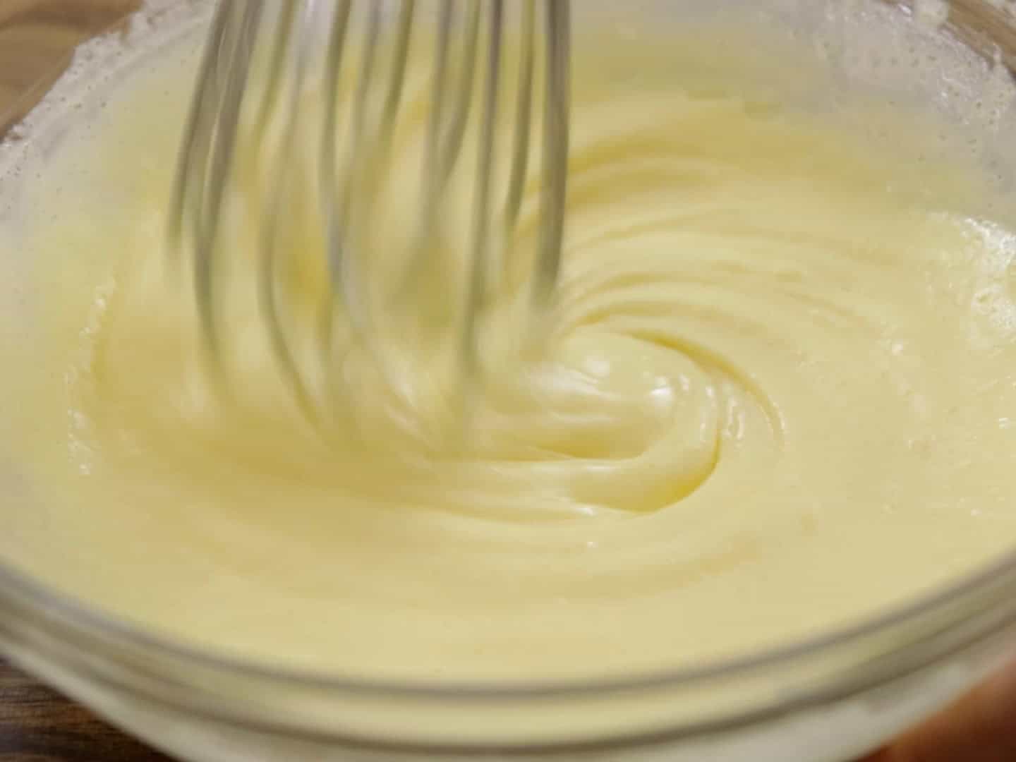 close-up of a metal whisk stirring a thick, pale yellow, creamy mixture in a clear glass bowl. The liquid has a smooth, custard-like consistency, forming soft swirls and ribbons as it’s whisked. The texture looks rich and glossy, suggesting a mixture like eggnog, custard, or a cream-based batter being blended until smooth. The focus is tight on the motion of the whisk and the creamy texture, with a warm, kitchen-style background barely visible.