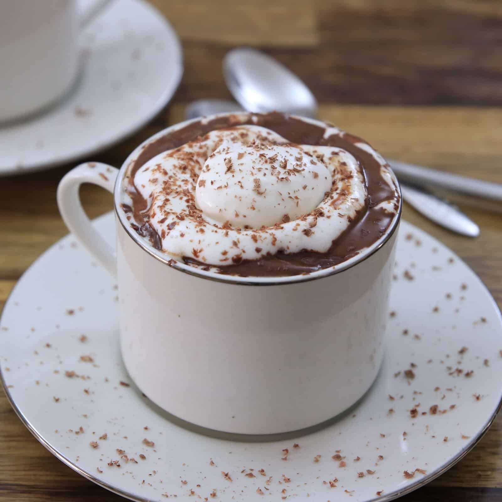A white cup filled with hot chocolate topped with whipped cream and chocolate shavings, sitting on a saucer. A spoon and another cup on a saucer are visible in the background on a wooden table.