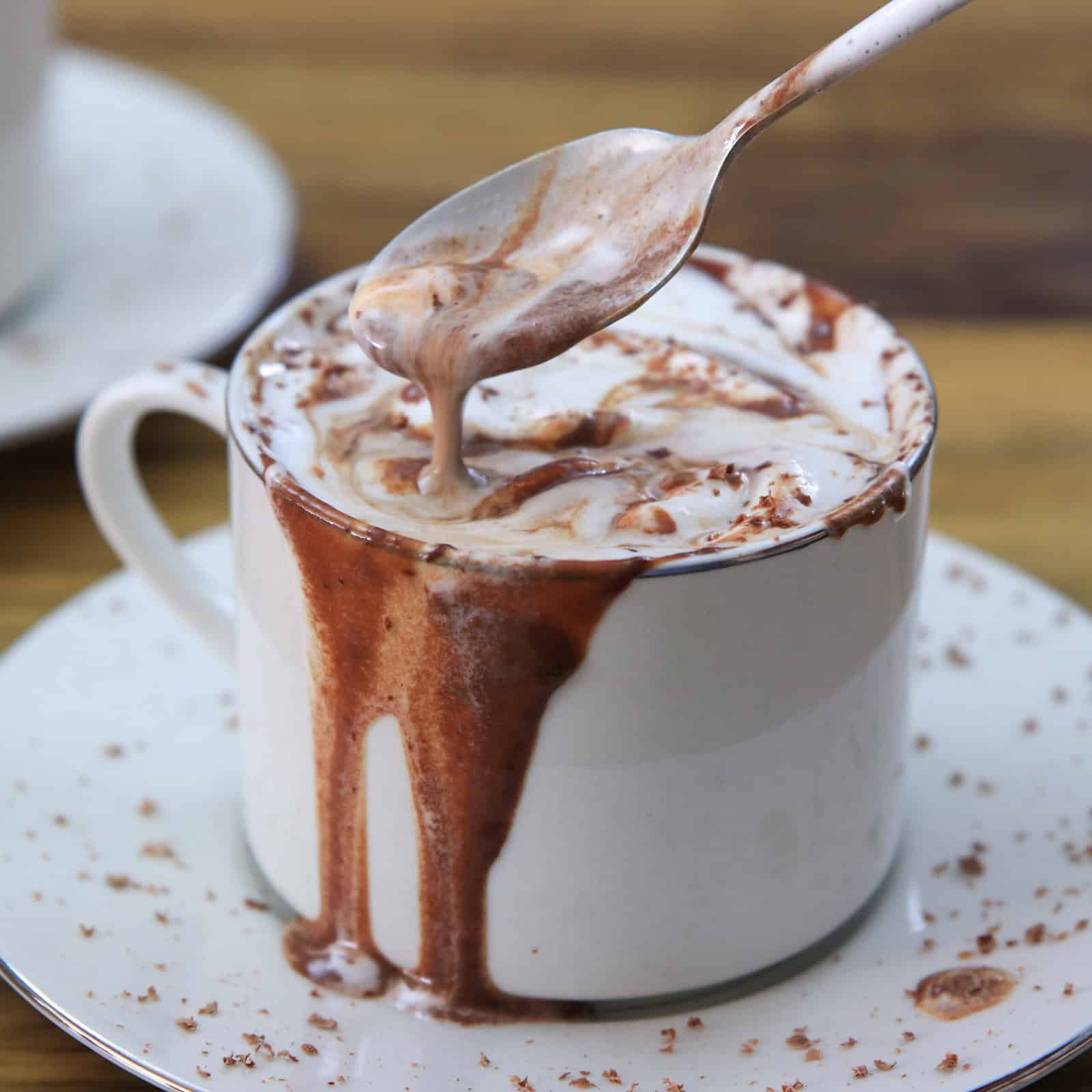 A white mug filled with frothy hot chocolate, overflowing and dripping chocolate down the sides, sits on a white saucer. A spoon with some hot chocolate is held above the mug.