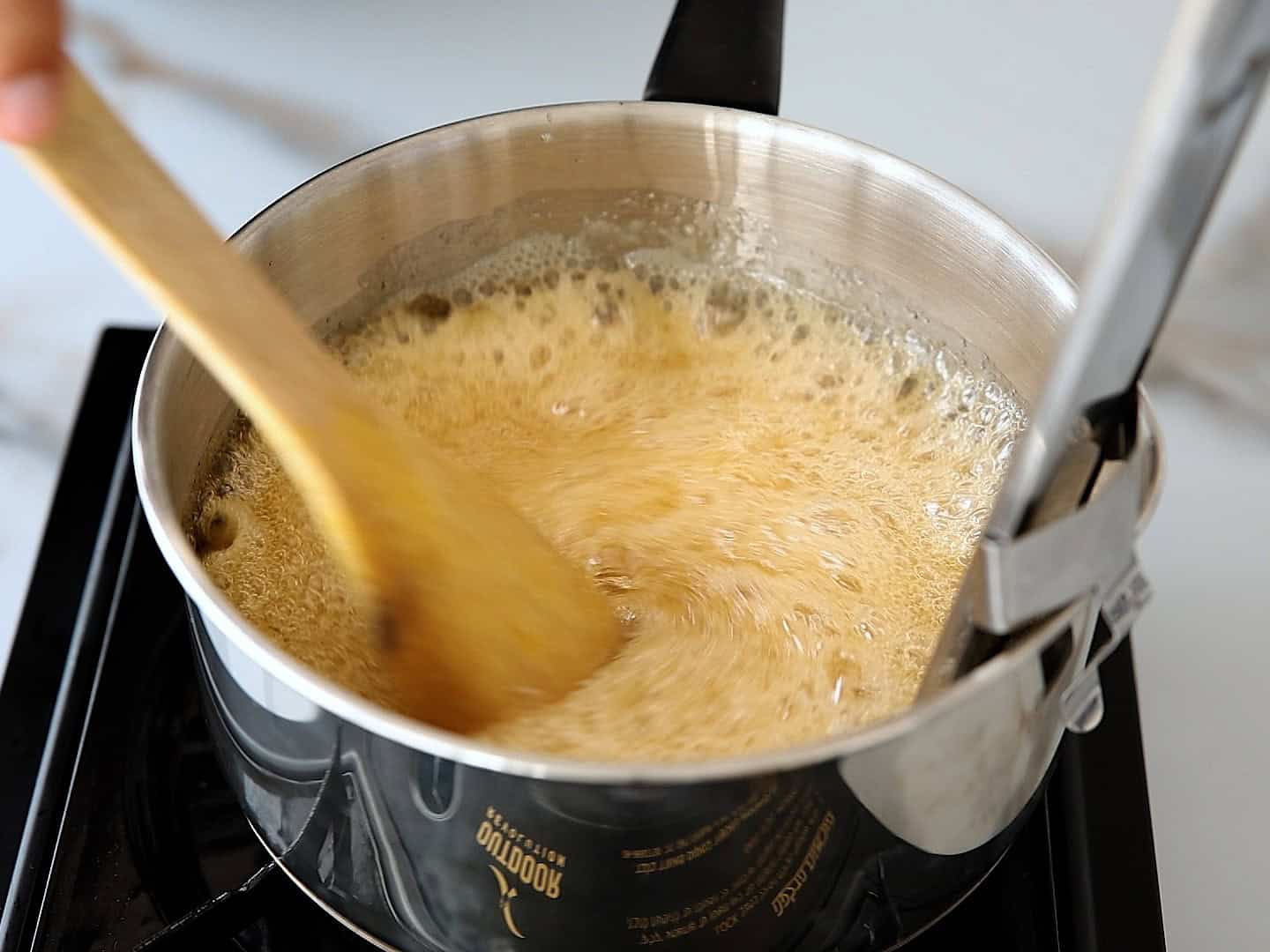 A person stirs bubbling caramel in a stainless steel pot on a stovetop with a wooden spoon.
