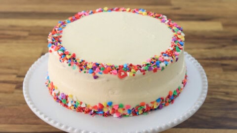 A round, frosted cake on a white plate, decorated with colorful sprinkles around the base and top edge, sitting on a wooden surface.