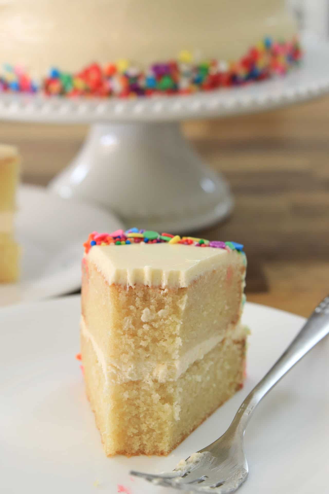 A slice of vanilla layer cake with white frosting and colorful sprinkles sits on a plate with a fork. The slice shows a bite taken out of the top. A whole decorated cake is blurred in the background.