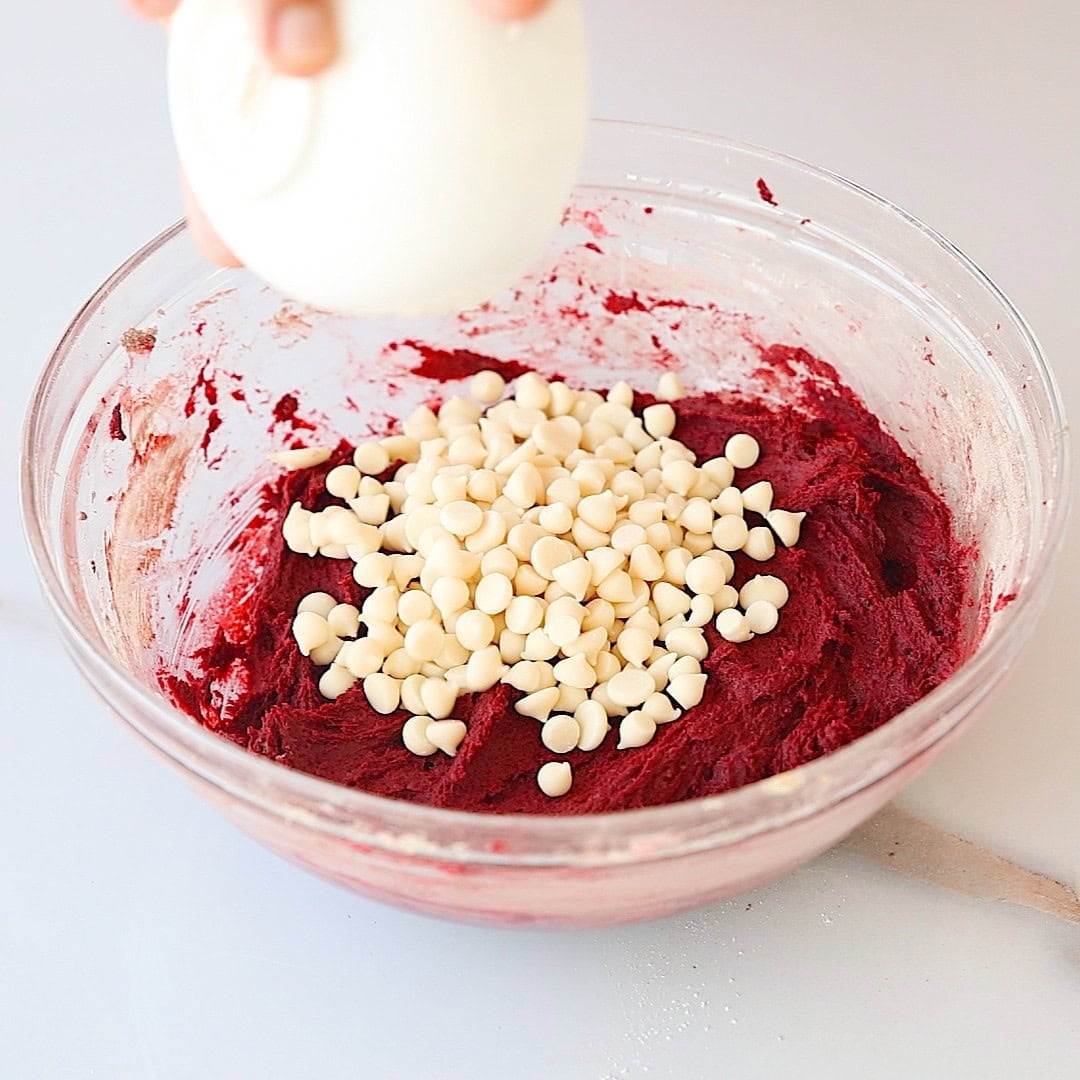 A glass bowl filled with red cookie dough and topped with white chocolate chips. A hand is holding a small white bowl above the mixture, as if pouring in more chips.