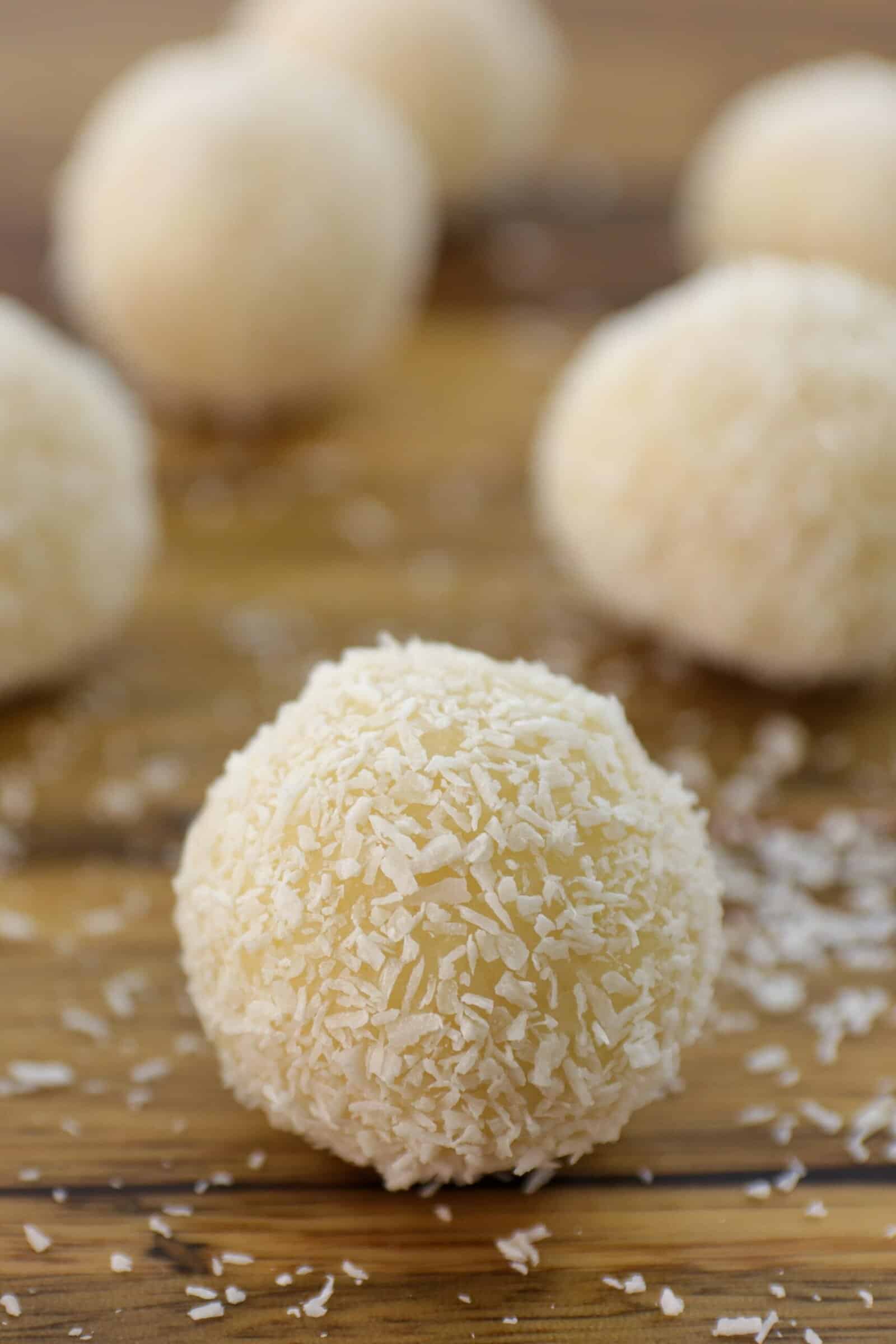 A close-up of round, white chocolate truffles coated in shredded coconut on a wooden surface, with more coconut balls blurred in the background.
